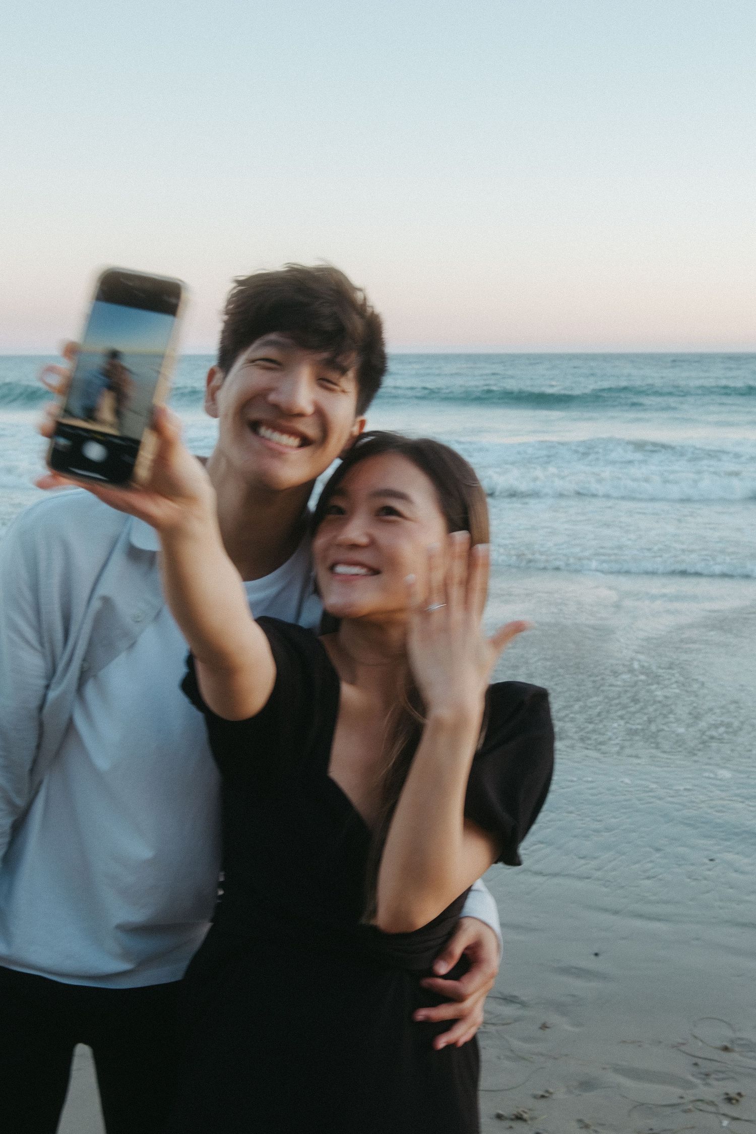 Happy engaged Asian couple taking selfie on beach at sunset, woman showing off engagement ring.