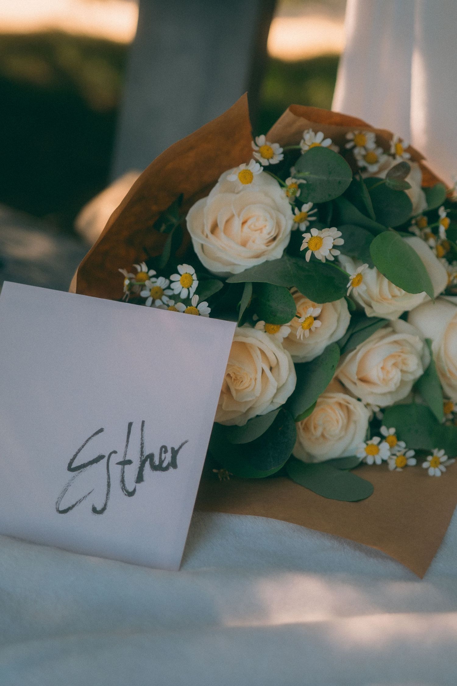 A bouquet of white roses and daisies with a card labeled Esther wrapped in brown paper.