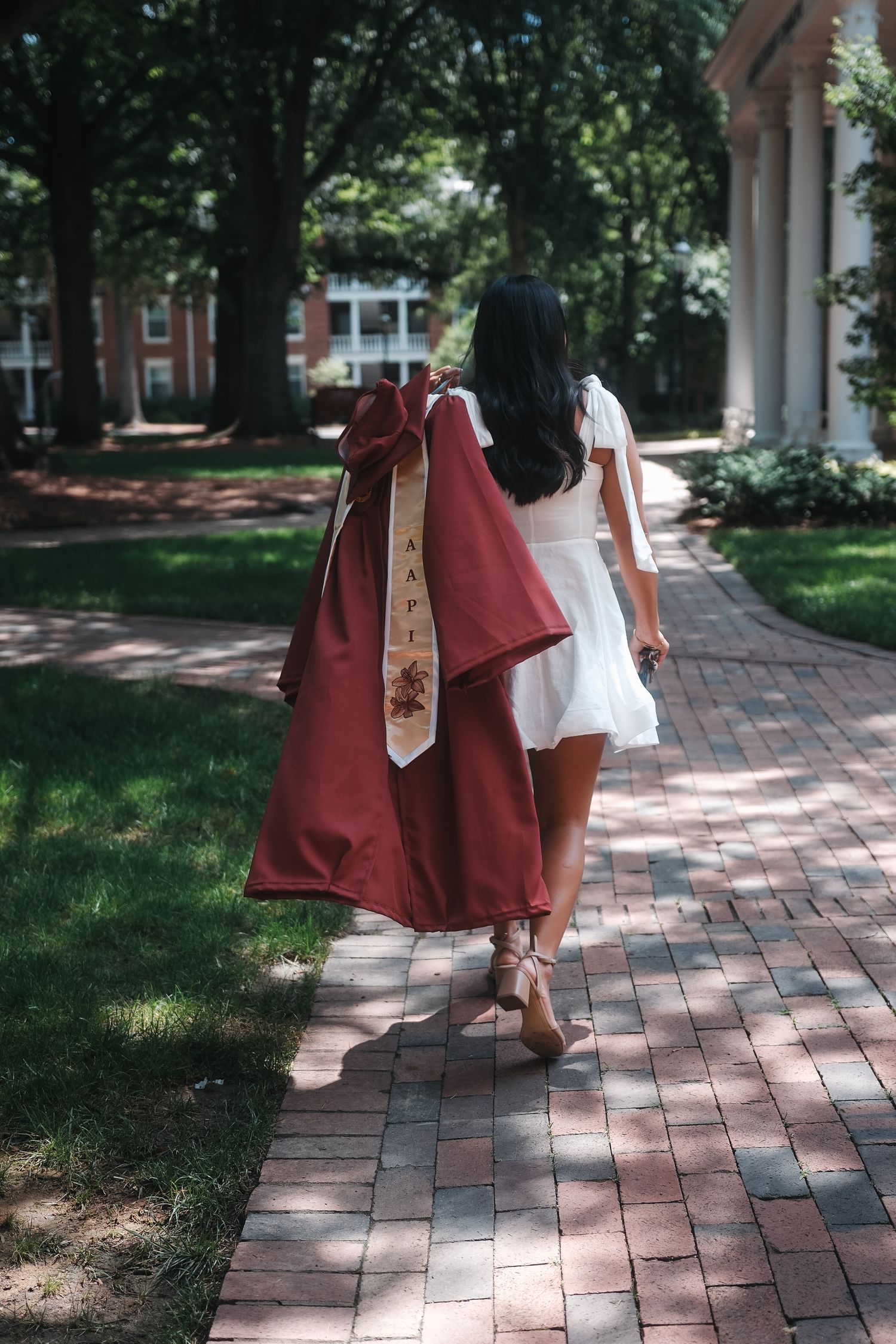 College graduate in maroon gown with decorative stole walks along brick pathway on campus.