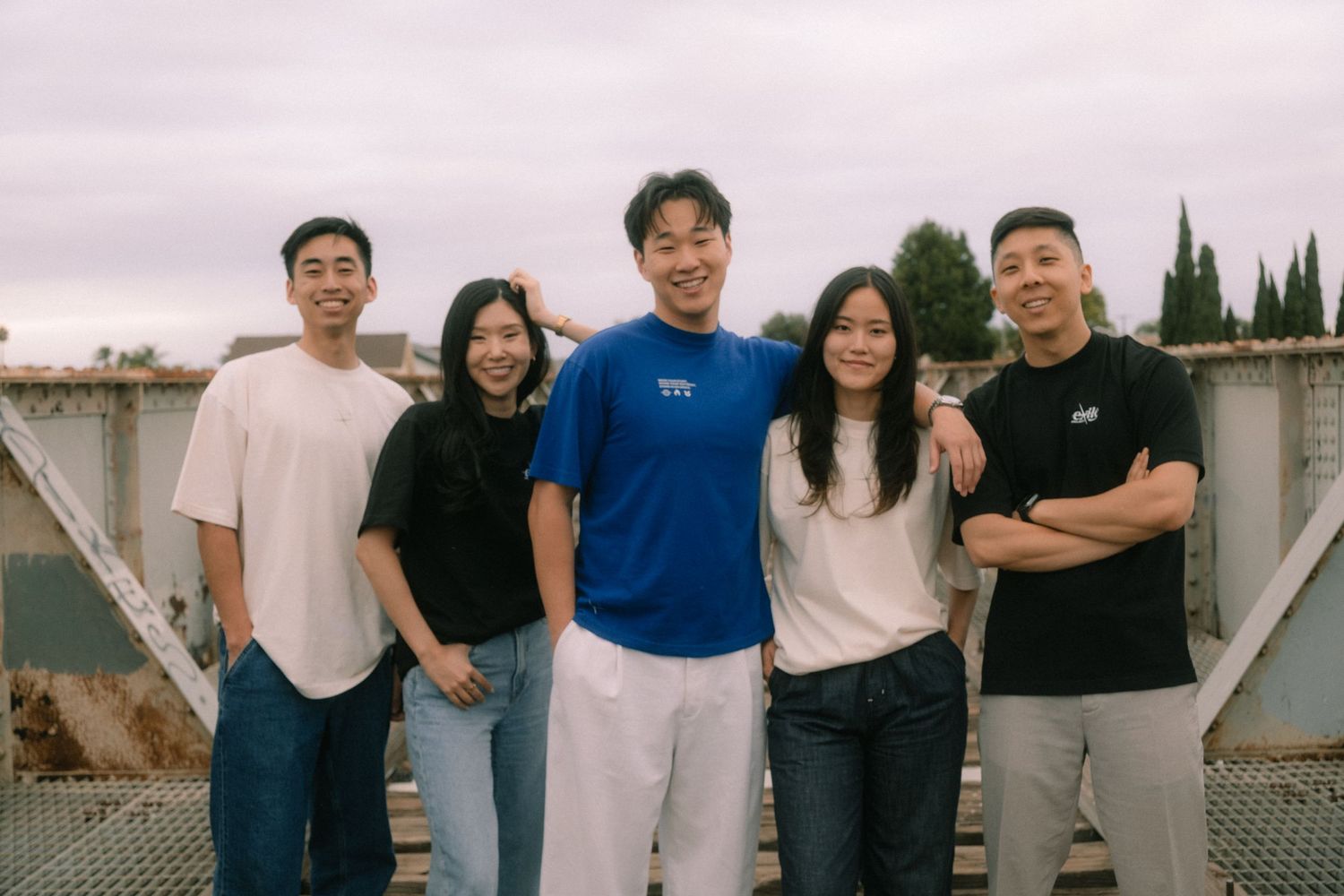 Group of five young Asian adults smiling together outdoors on a cloudy day.