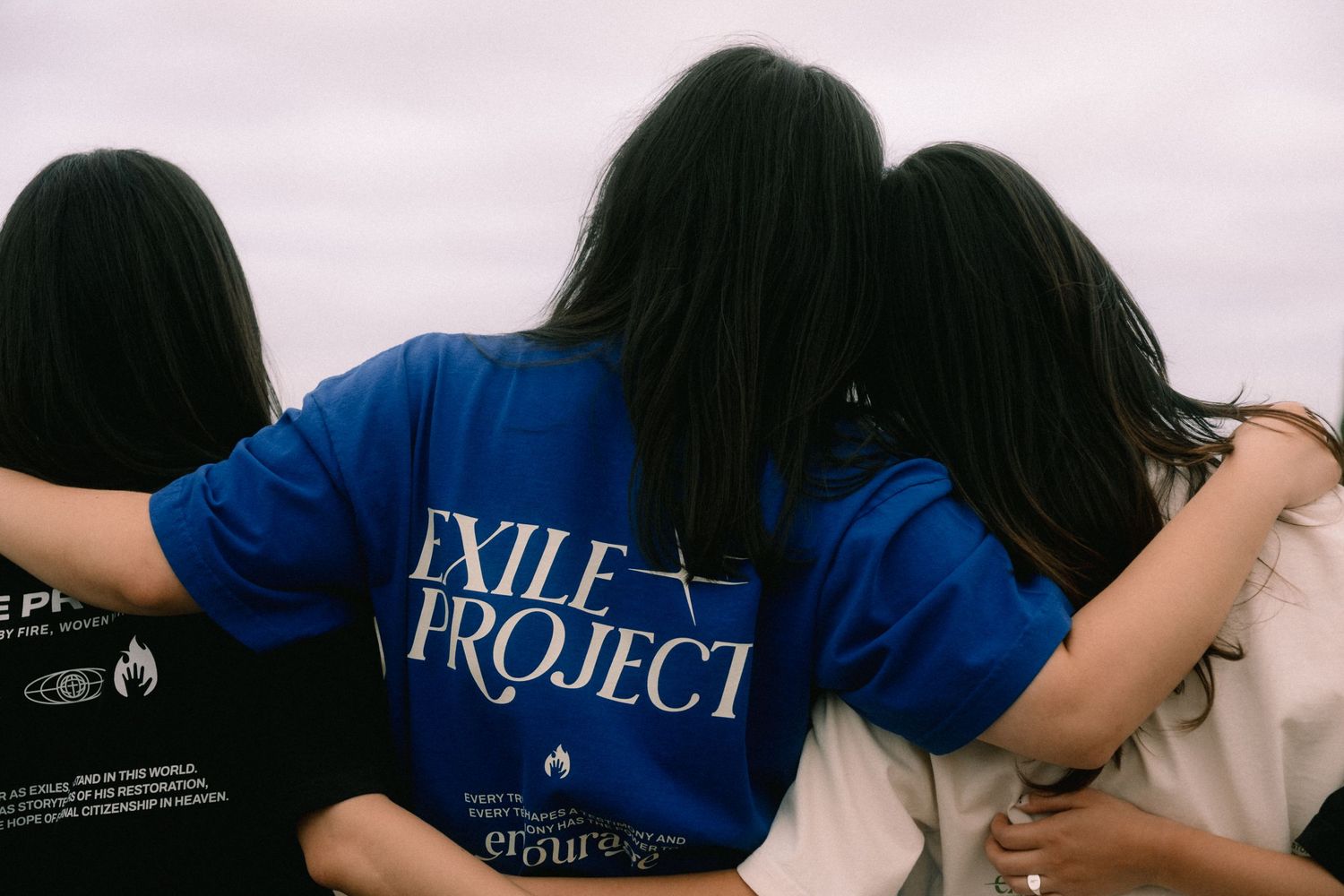 Three young women with dark hair embrace, wearing Exile Project blue and white t-shirts, photographed from behind outdoors.