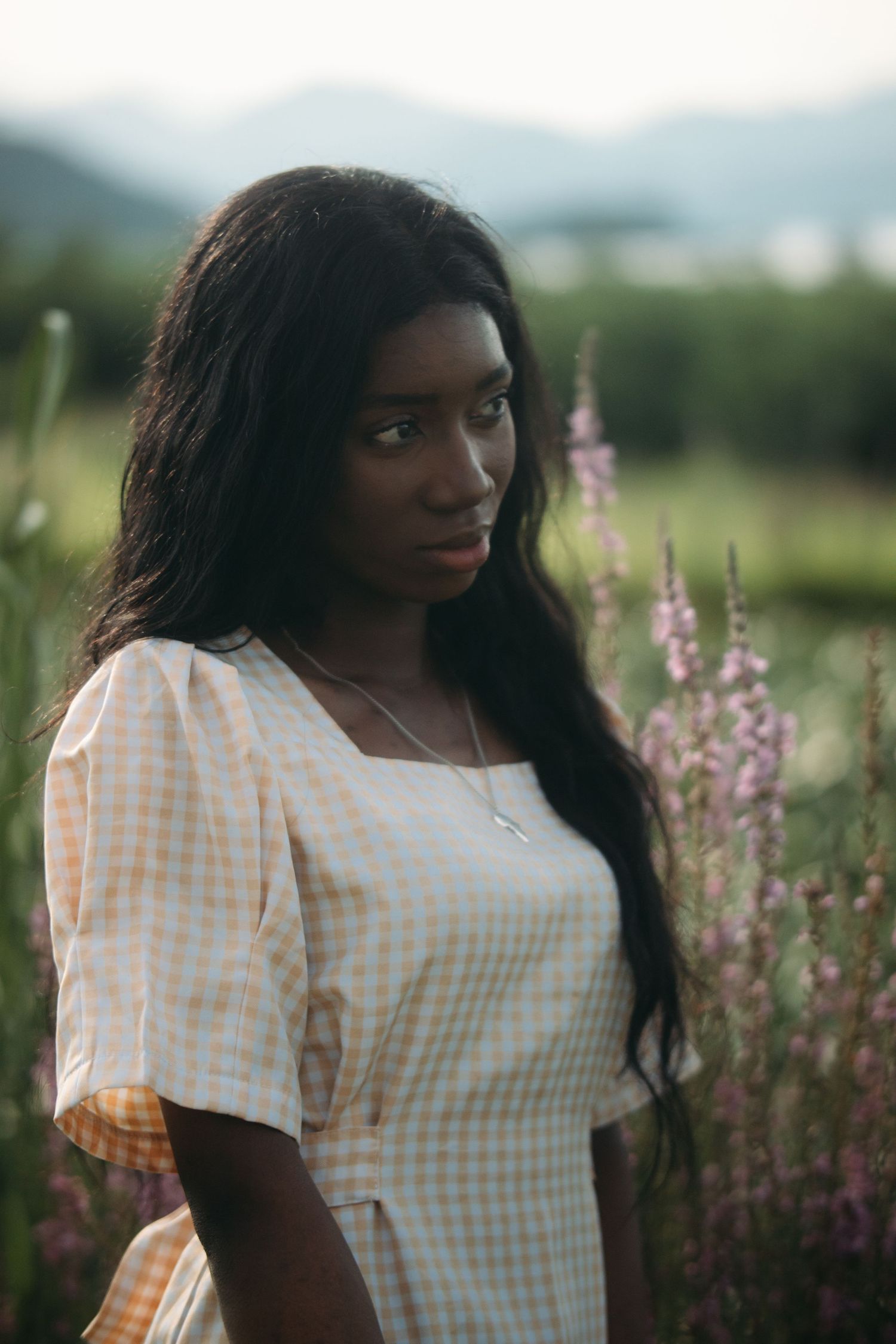 Young Black woman in yellow gingham dress stands among purple wildflowers with mountains behind.