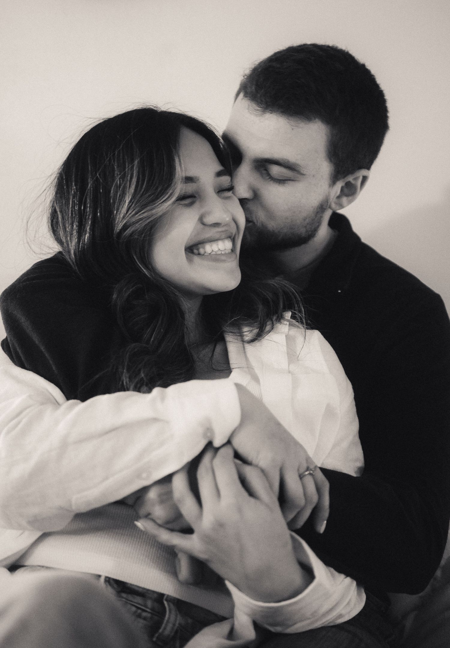 Black and white photo of a smiling woman being hugged and kissed by a man, capturing a tender romantic moment.