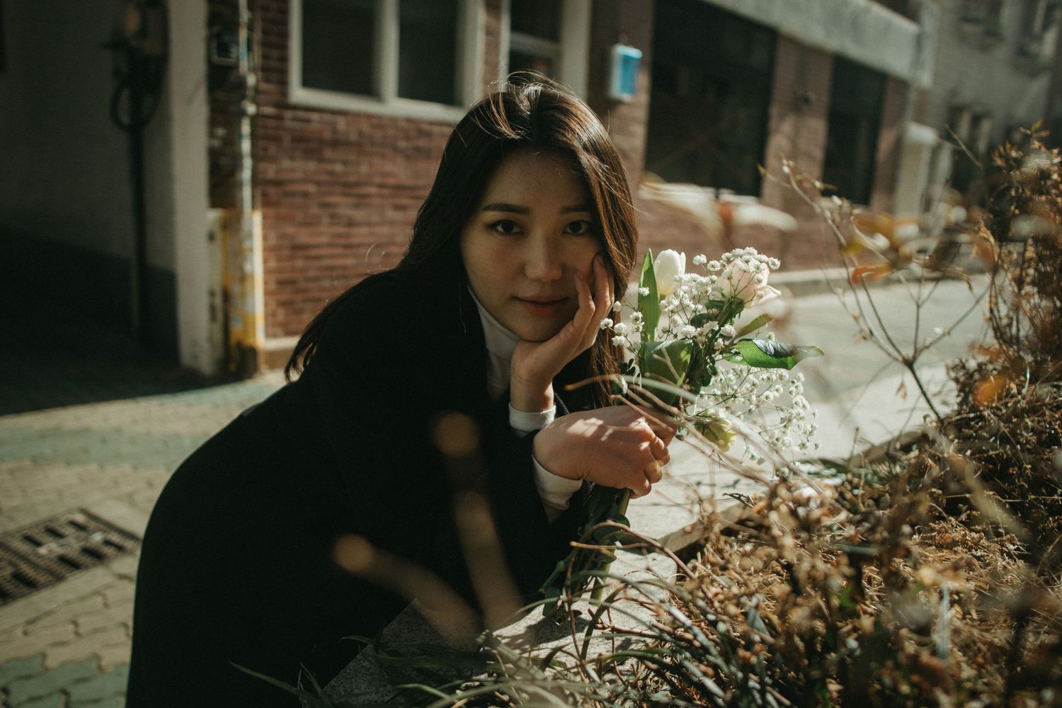 Young woman crouching outdoors holding white flowers, posing near dried plants in sunlight.