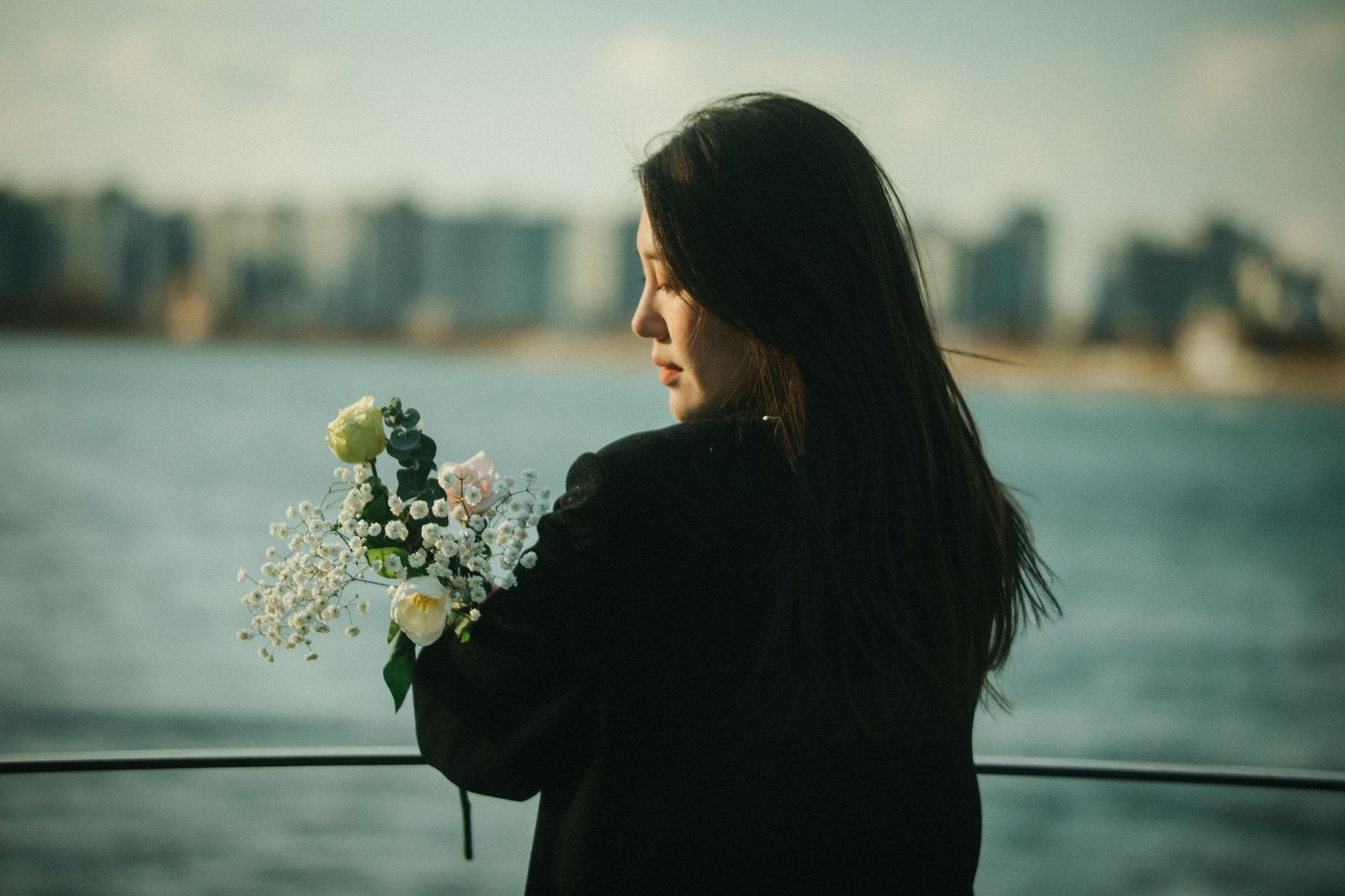 Young Asian woman in black holding a white flower bouquet by a waterfront with a blurred city skyline.