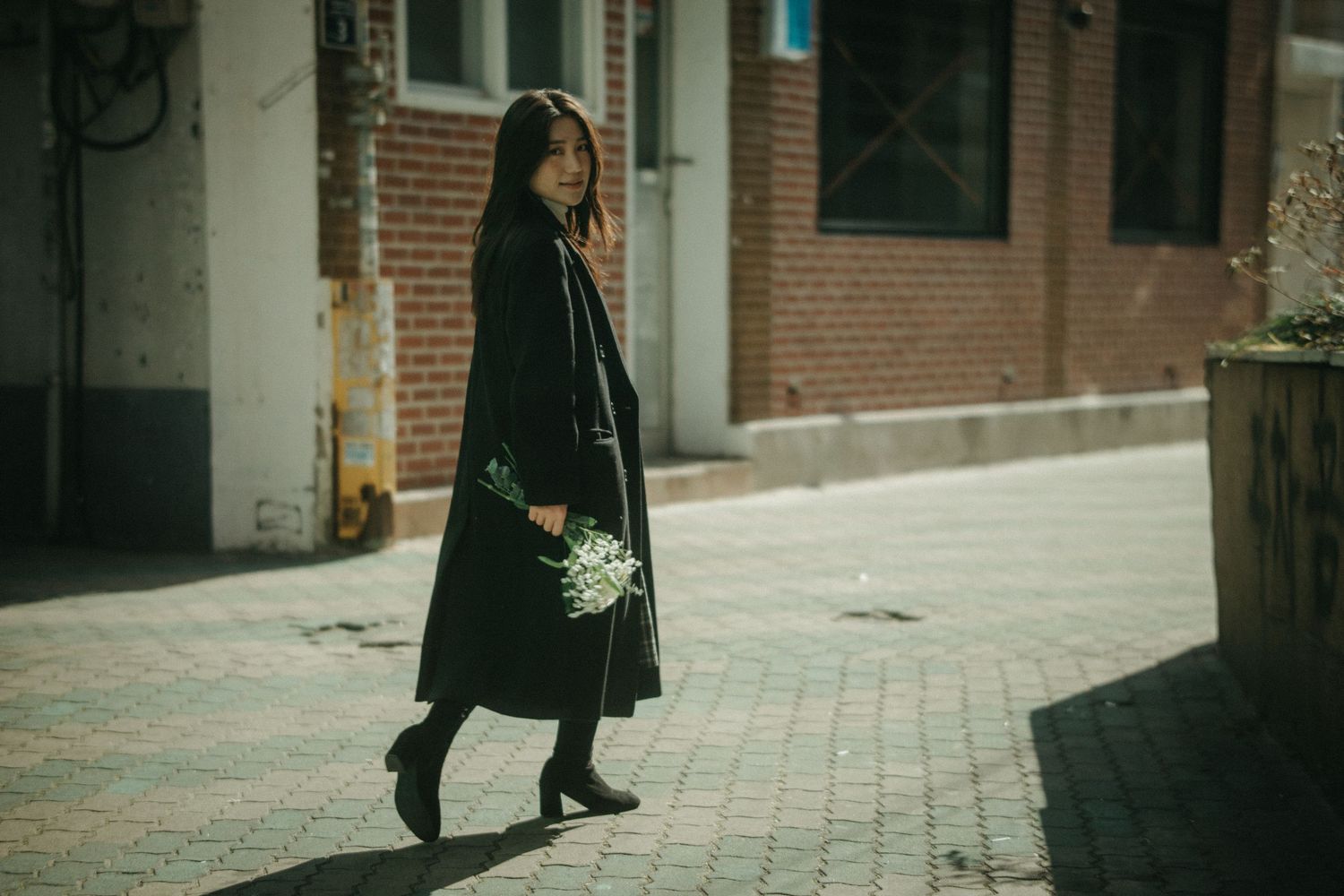 Woman in black coat walking on brick sidewalk holding white flowers near brick building.