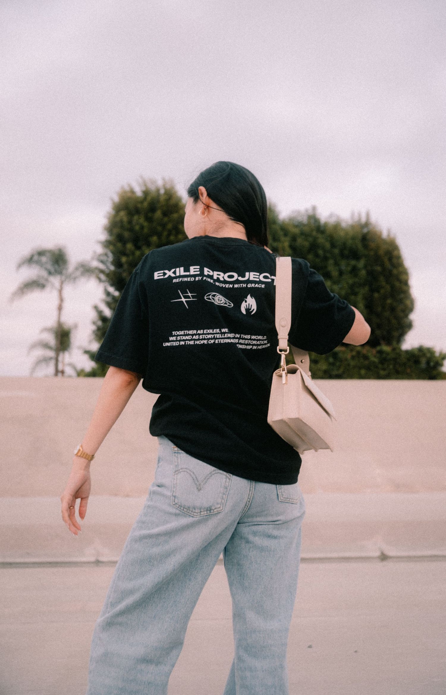 Person wearing black Exile Project graphic tee, light wash jeans, and carrying a beige bag, photographed from behind outdoors.