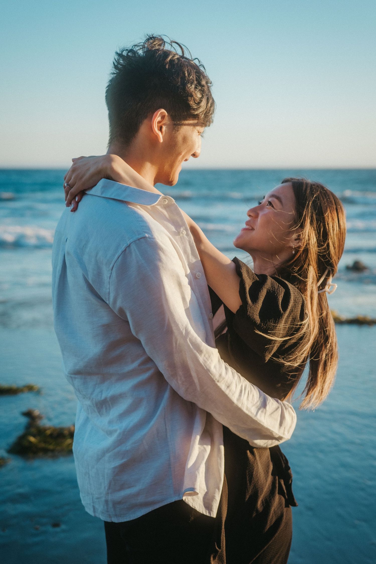Happy couple embracing and laughing together on a sunny beach with ocean waves in the background.
