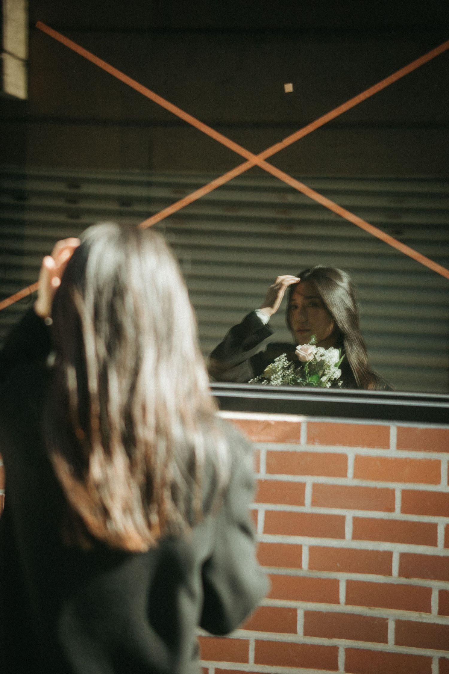 Asian woman adjusts hair while viewing reflection in X-taped window holding white flower bouquet.