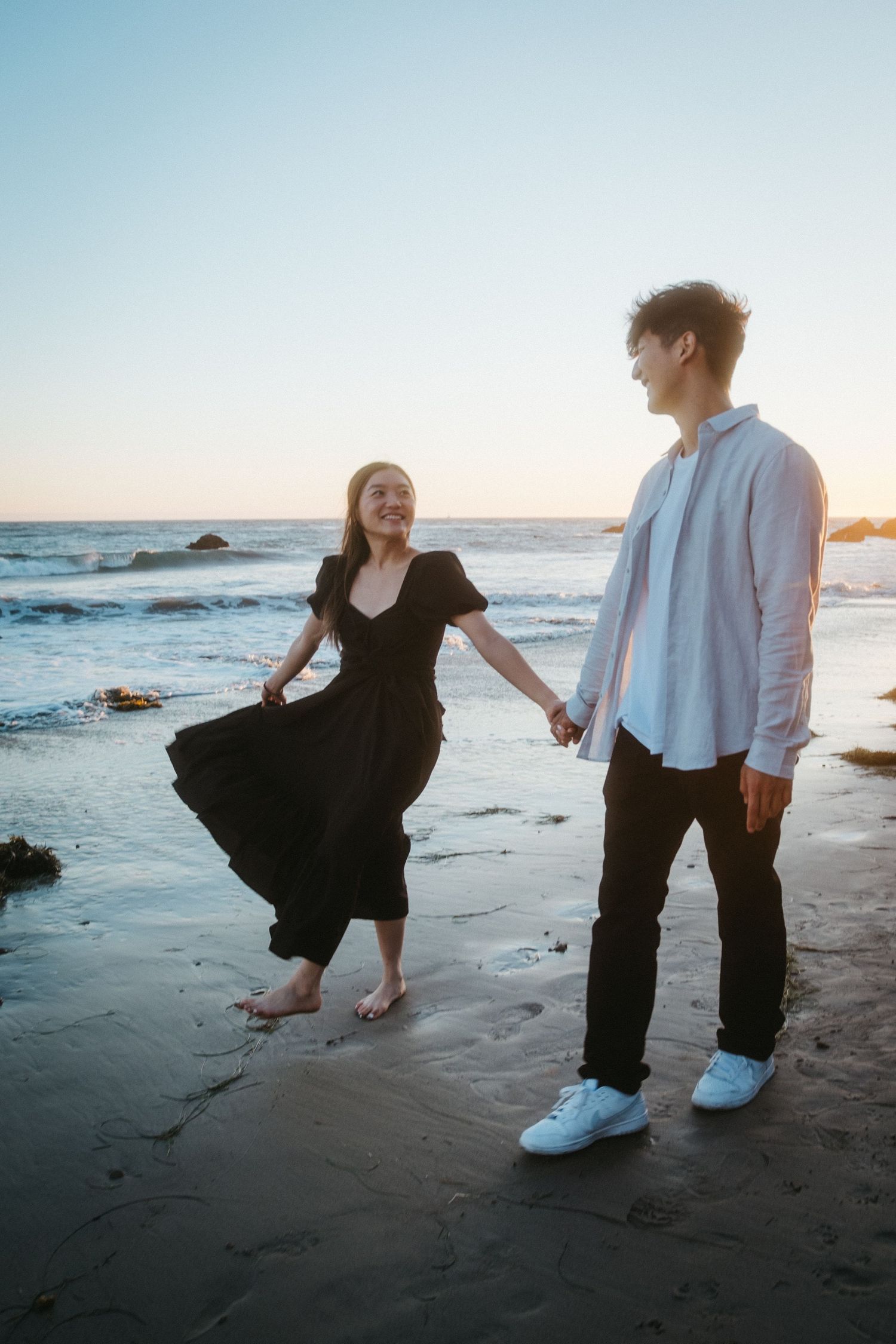 Joyful couple holding hands on a beach at sunset, woman in black dress laughing while facing her partner.