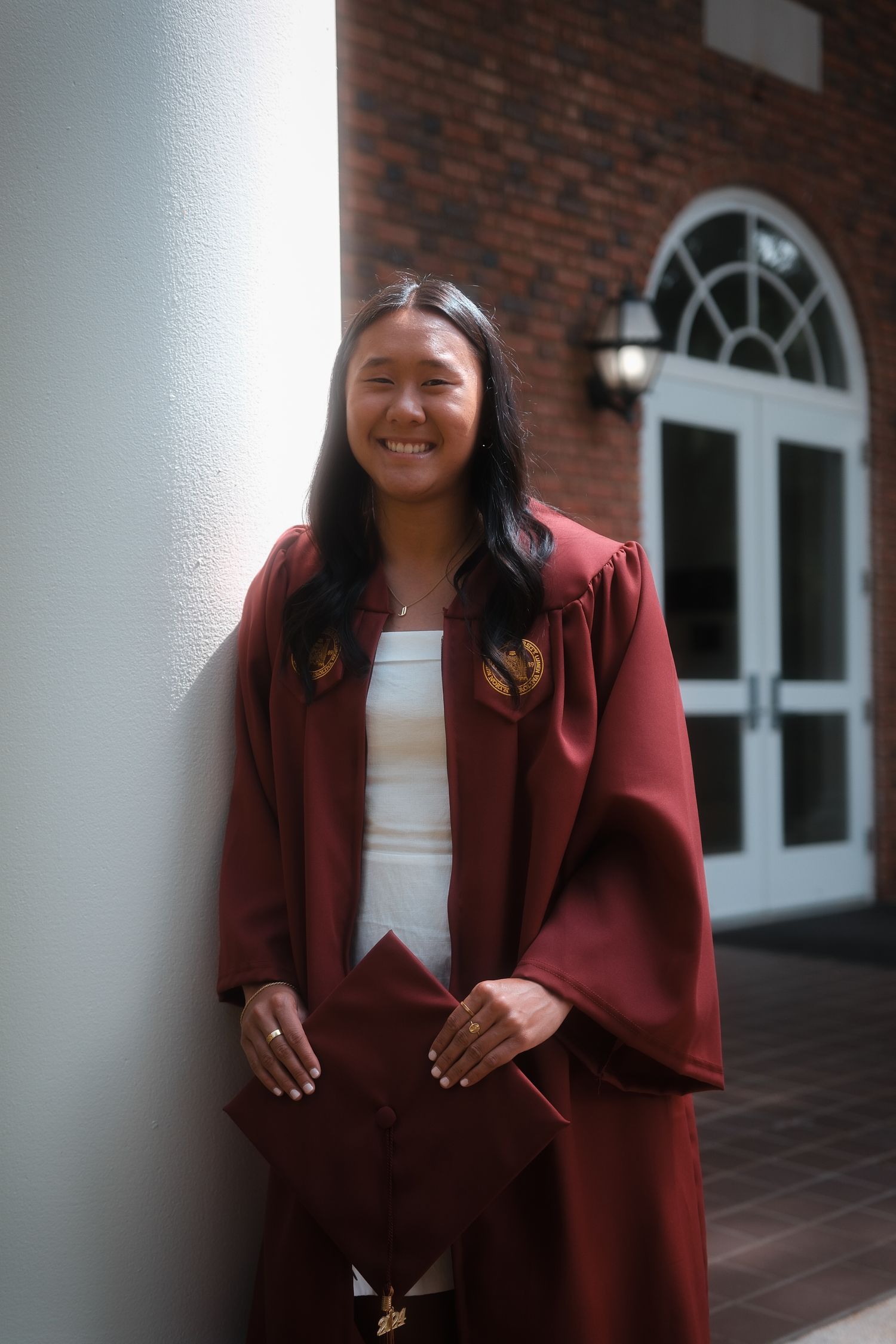 Happy graduate in maroon gown leans against white pillar holding cap at Elon University campus.