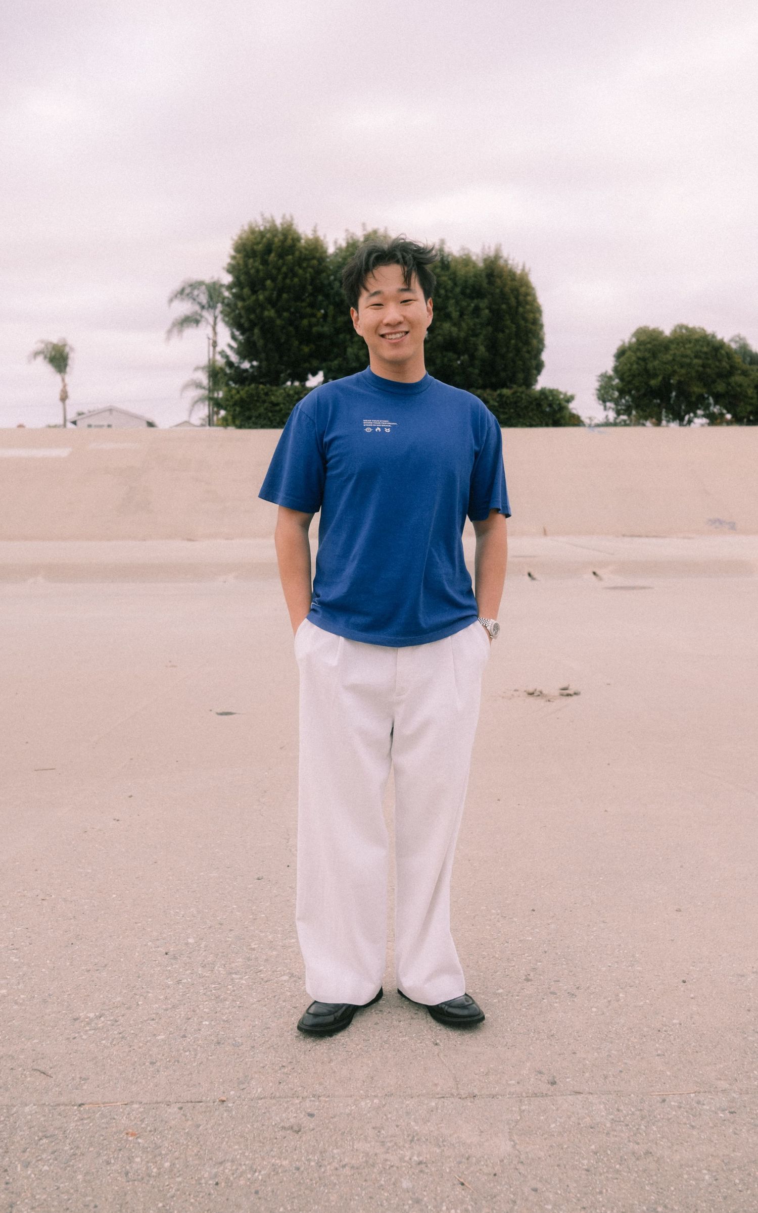 Smiling young man in blue graphic tee and white wide-leg pants standing on concrete outdoors.