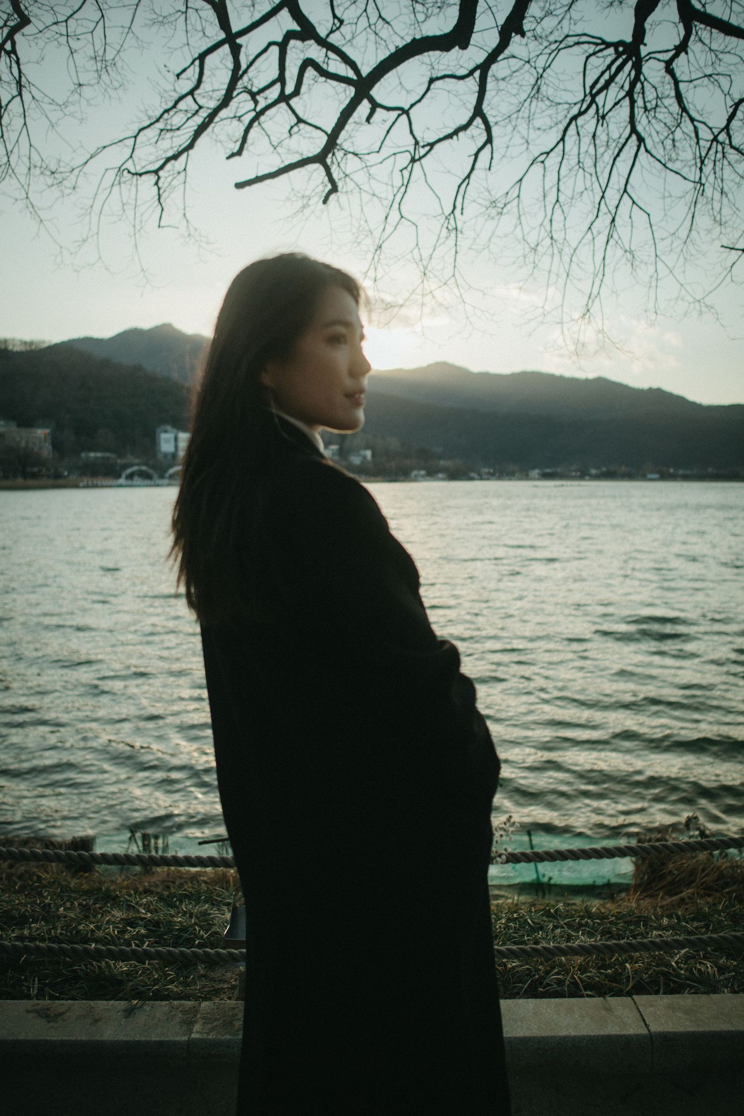 Young woman in black coat stands by lake under bare winter branches at glowing sunset.