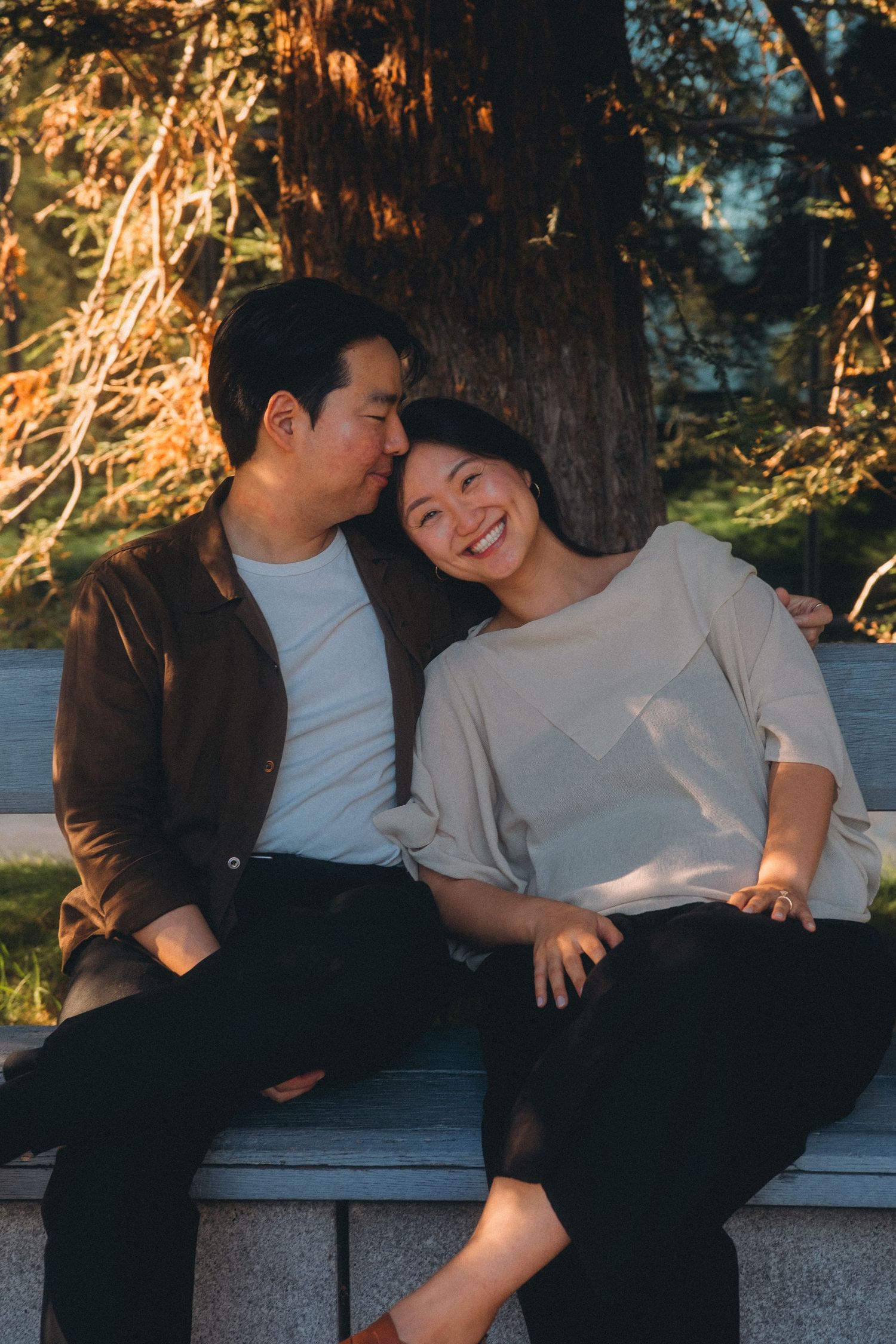 Happy Asian couple sitting together on a bench under a tree in warm golden hour sunlight.