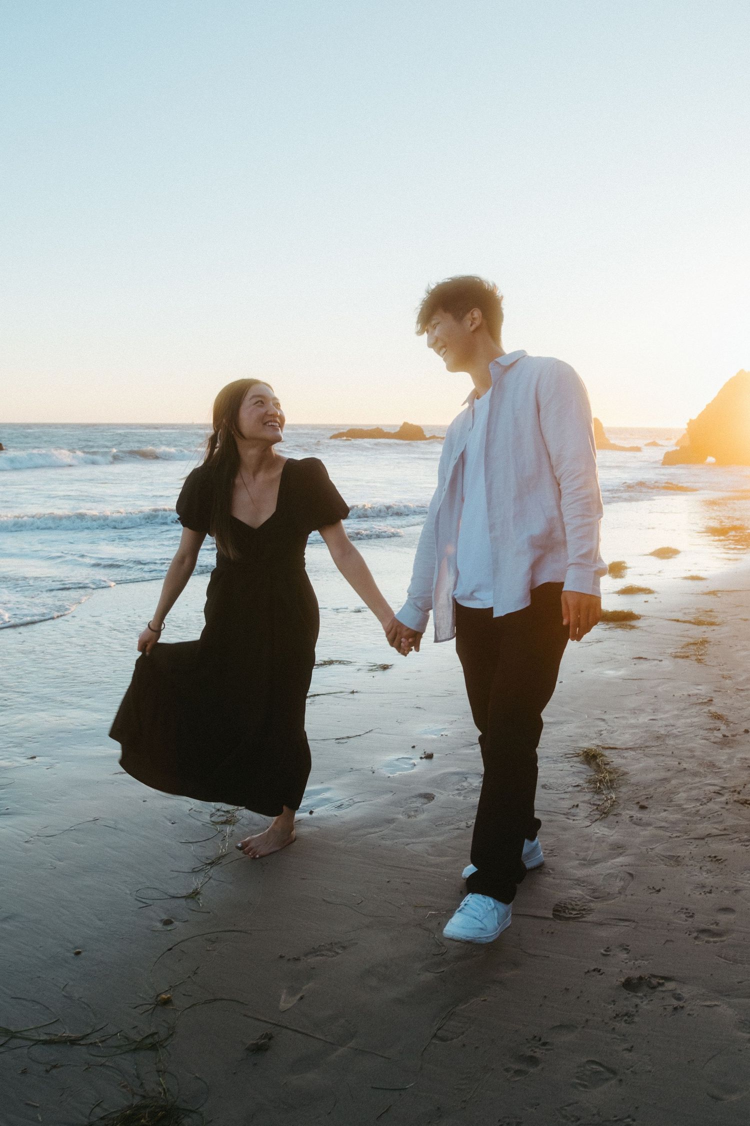Smiling couple holding hands on a beach at golden hour, woman in black dress and man in white shirt.