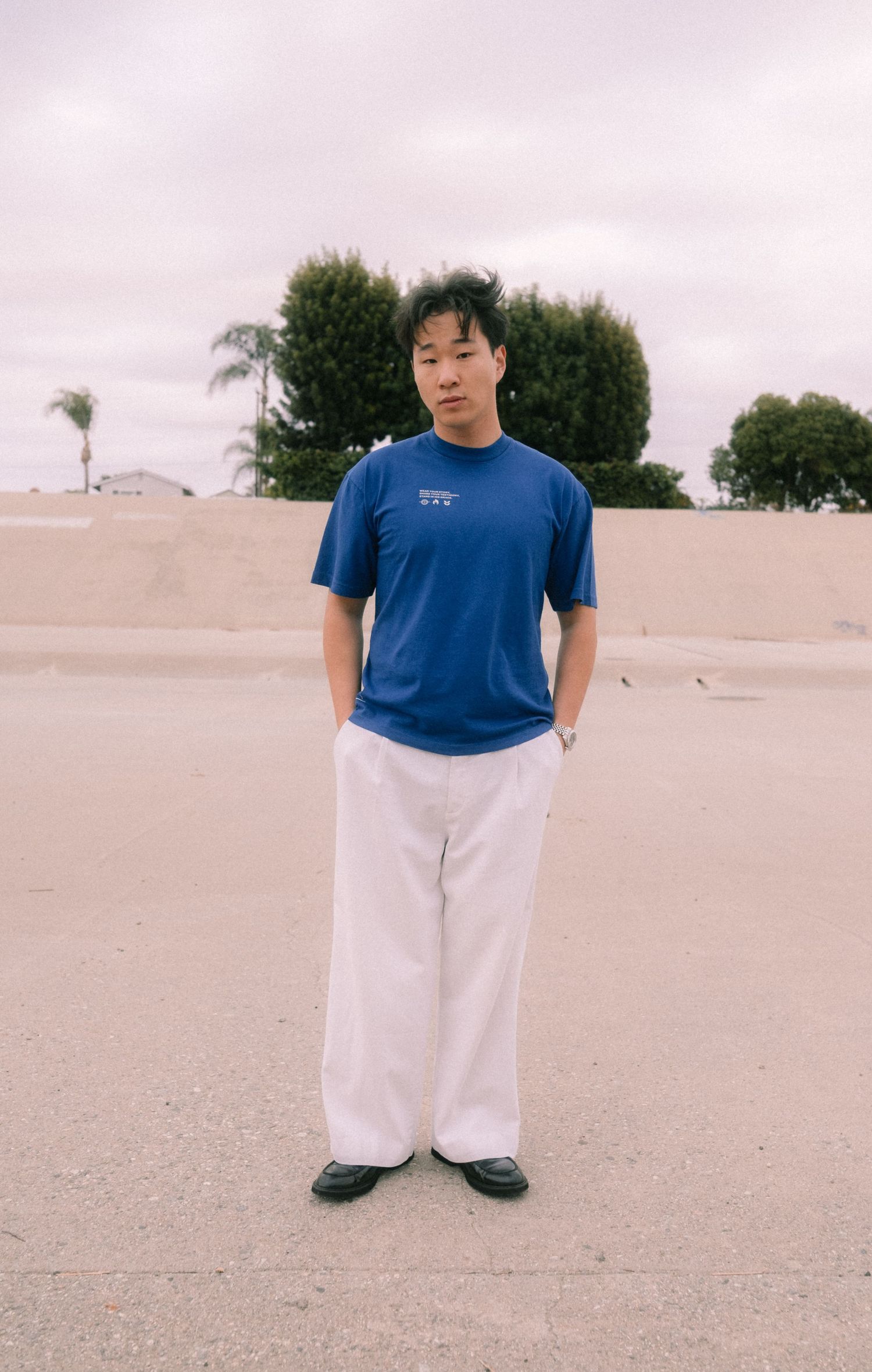 Young Asian man in blue graphic tee and white wide-leg pants standing on sandy ground.