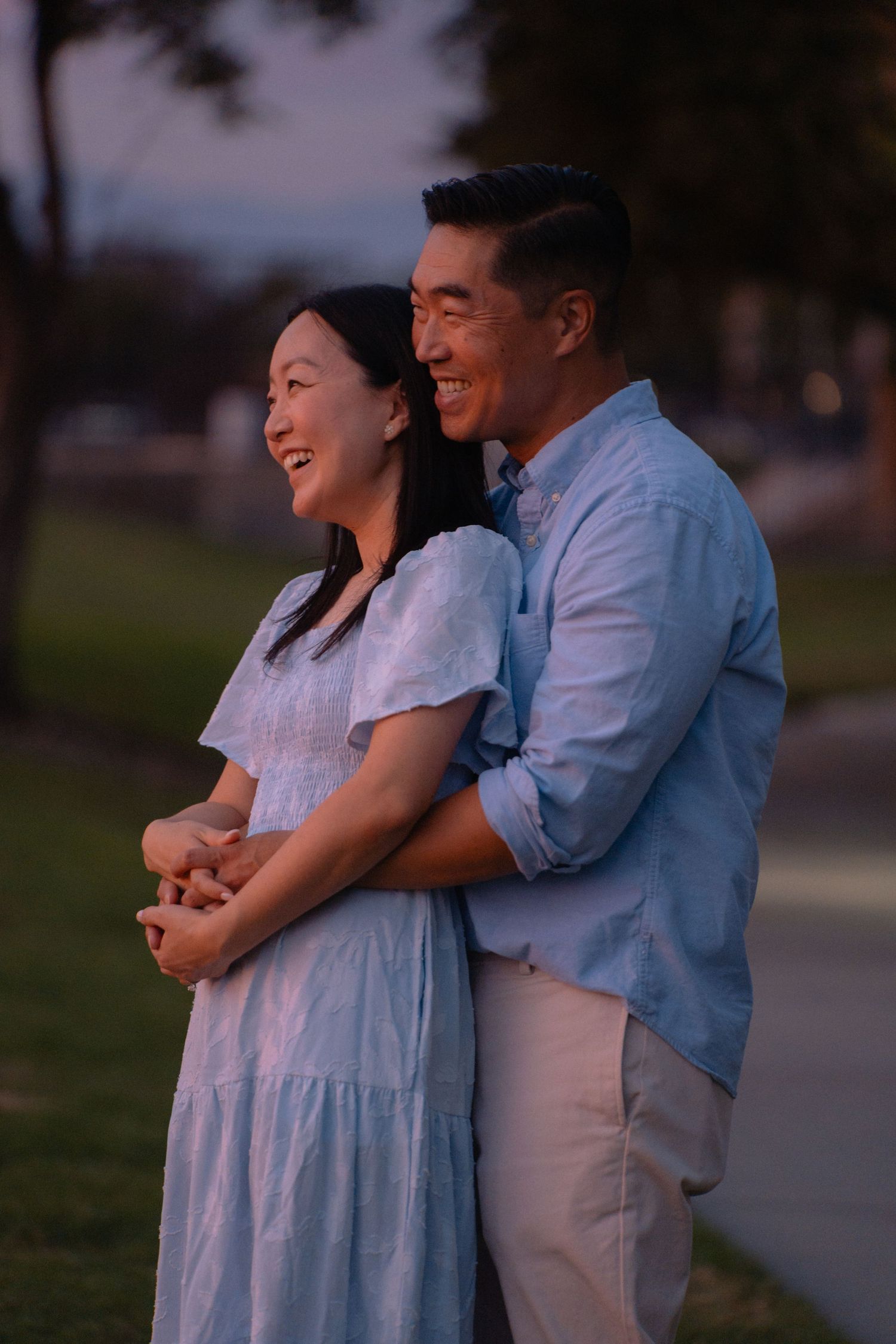 Happy Asian couple embracing outdoors at dusk, woman in blue dress and man in light blue shirt smiling together.