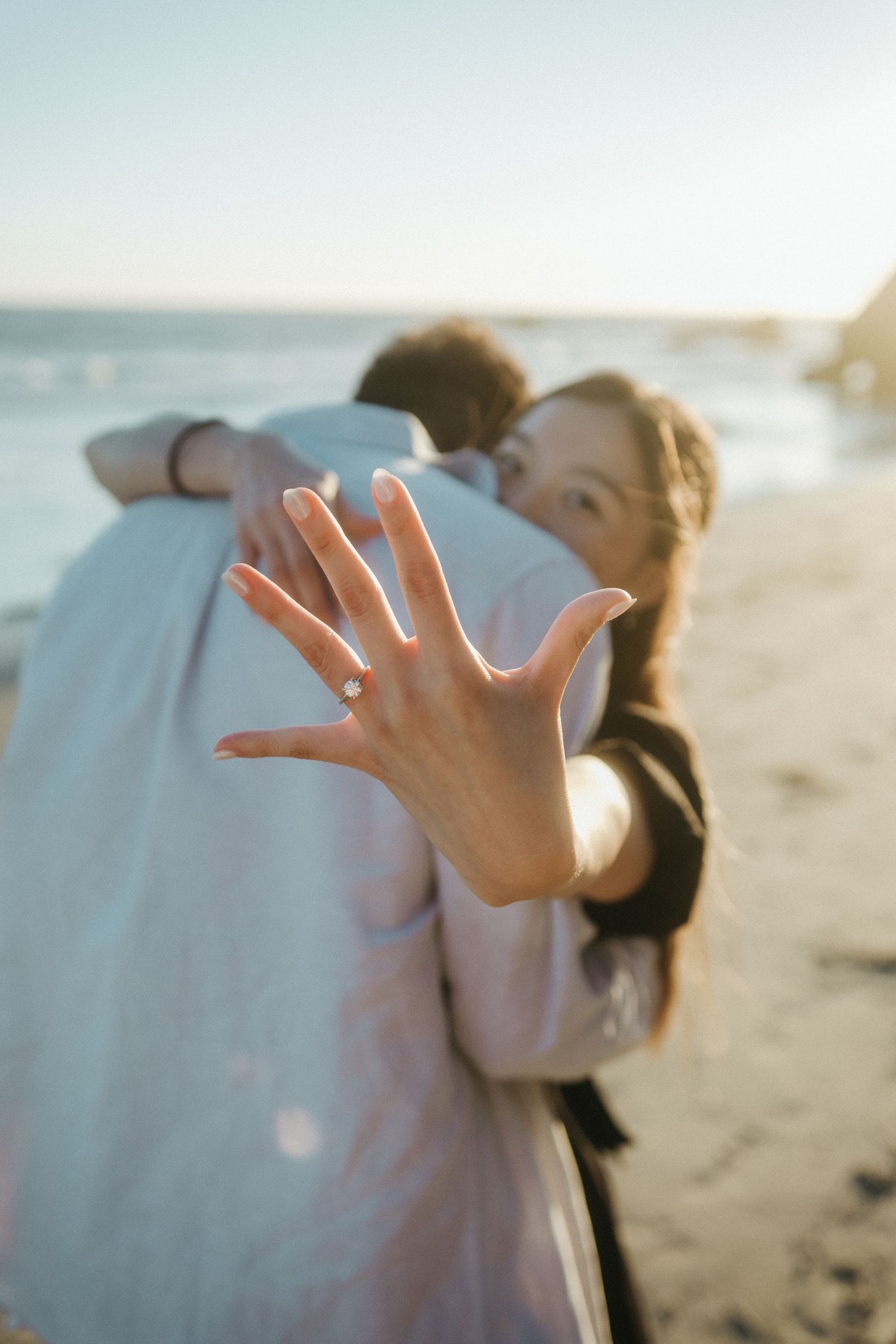 Newly engaged woman showing diamond ring on beach while embracing her partner at sunset.
