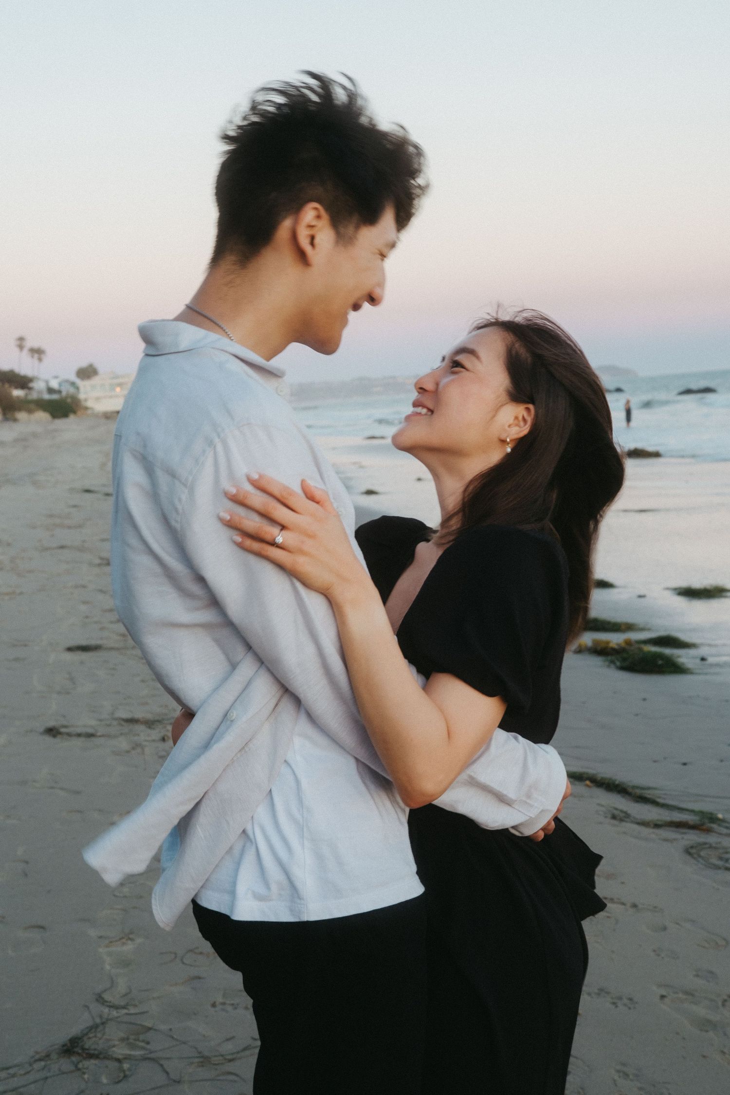 Joyful newly engaged Asian couple embracing and smiling at each other on beach at dusk.