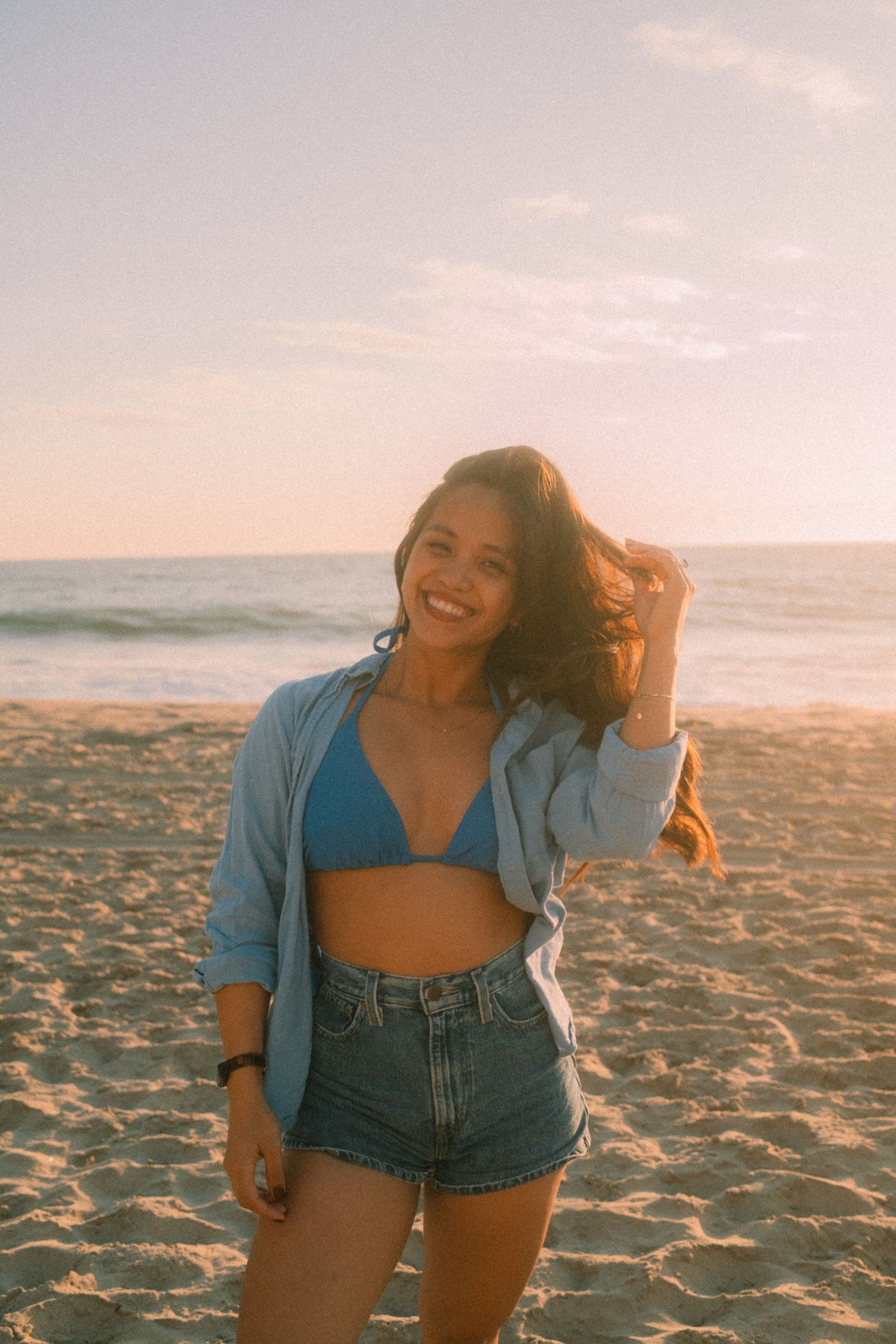 Joyful young woman in blue bikini top and jean shorts laughs while holding her hair on a beach at sunset.