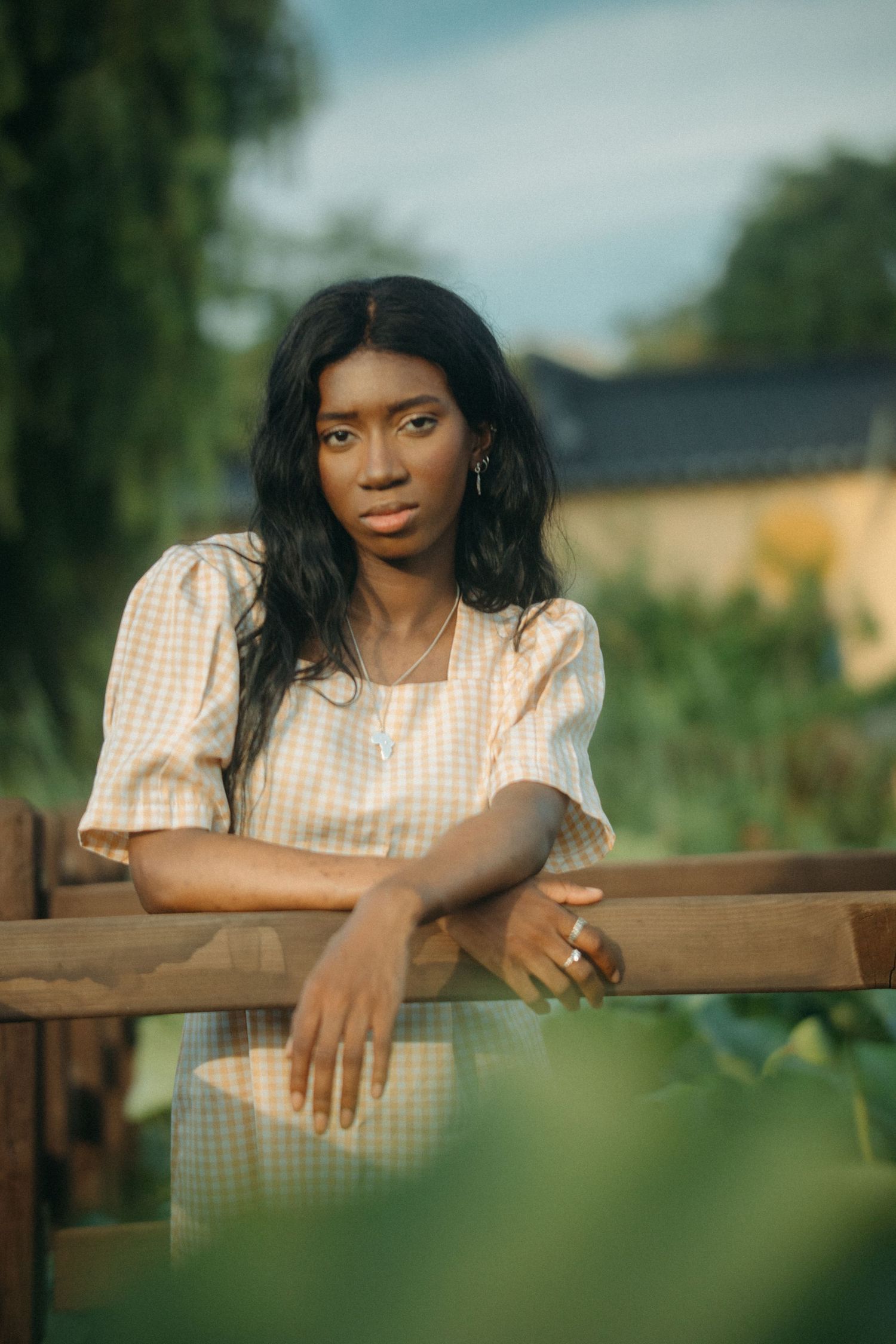 A young Black woman in a gingham dress leans on a wooden fence surrounded by lush garden greenery.