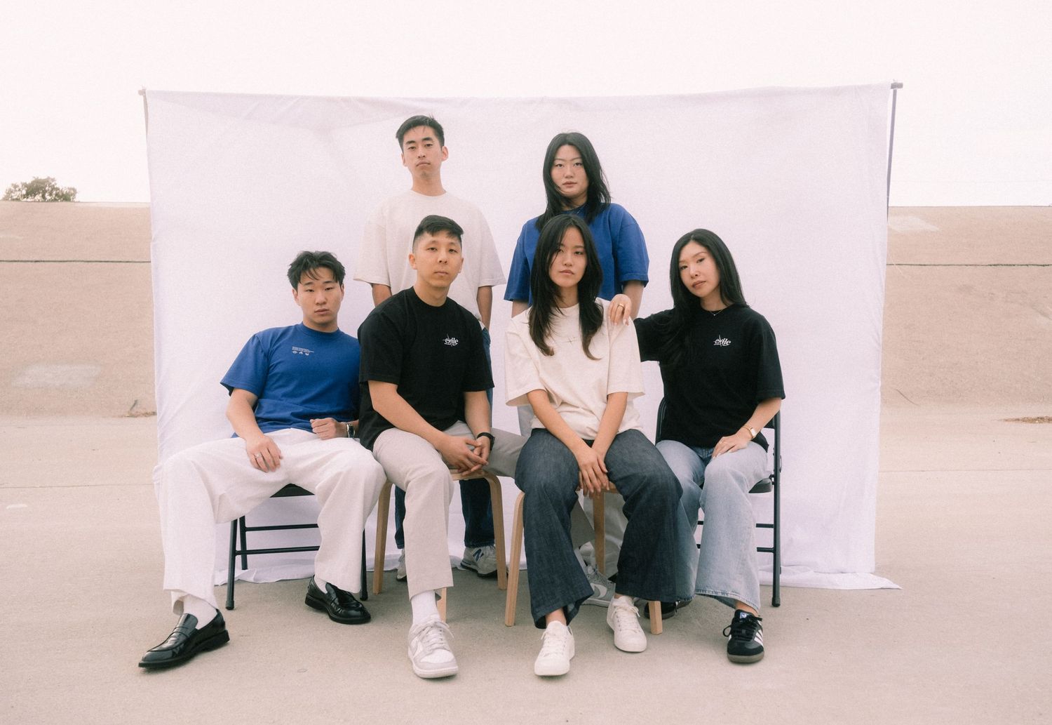 Six young adults pose together outdoors against a white backdrop, dressed in casual streetwear.