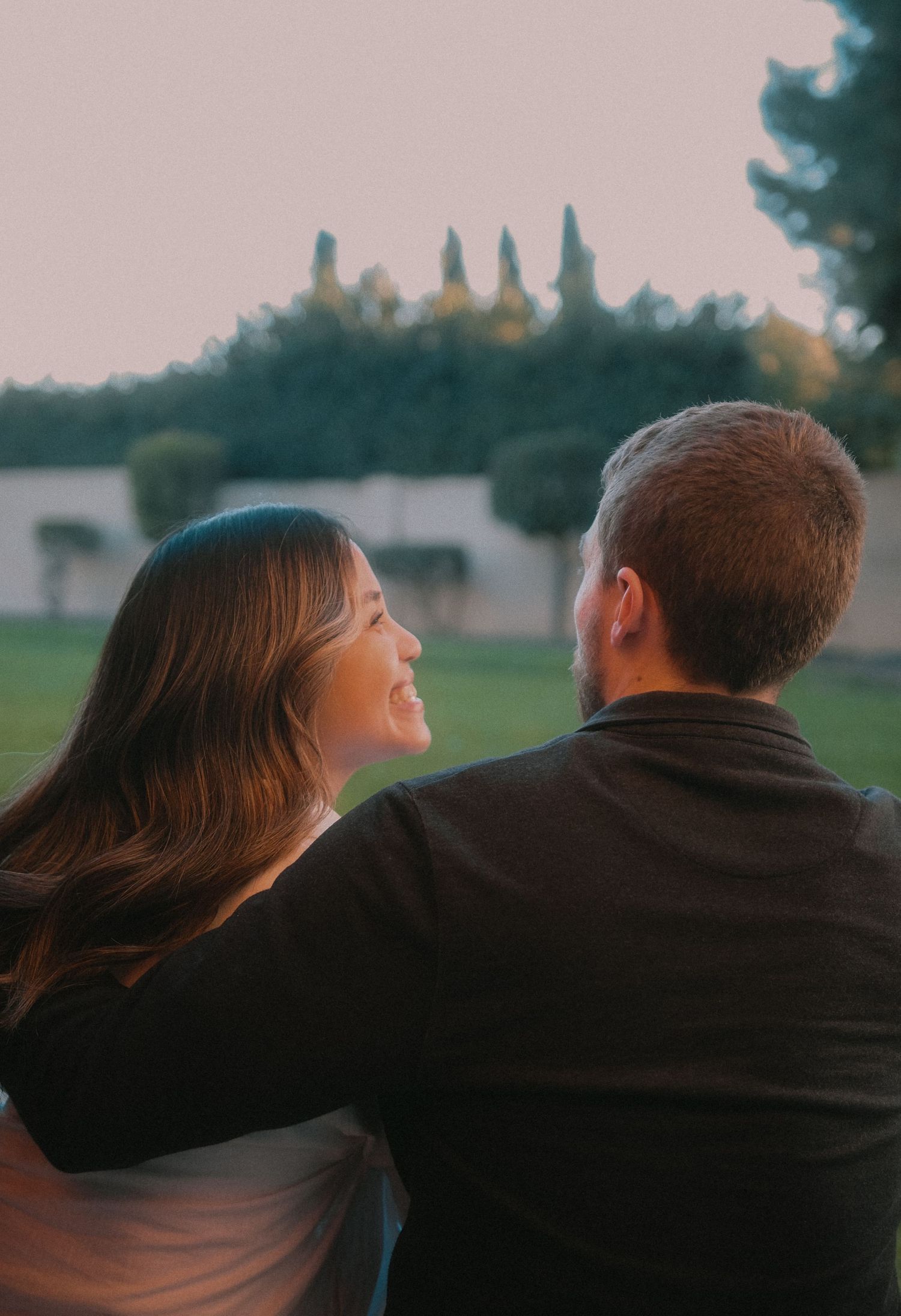 Couple sitting together outdoors at sunset, woman smiling, man with arm around her, green trees in background.