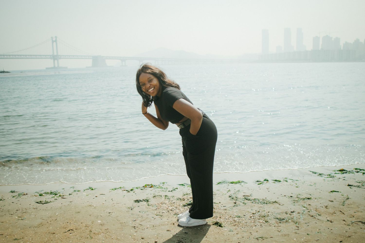 Woman in black outfit posing playfully on a beach with a bridge and city skyline in background.