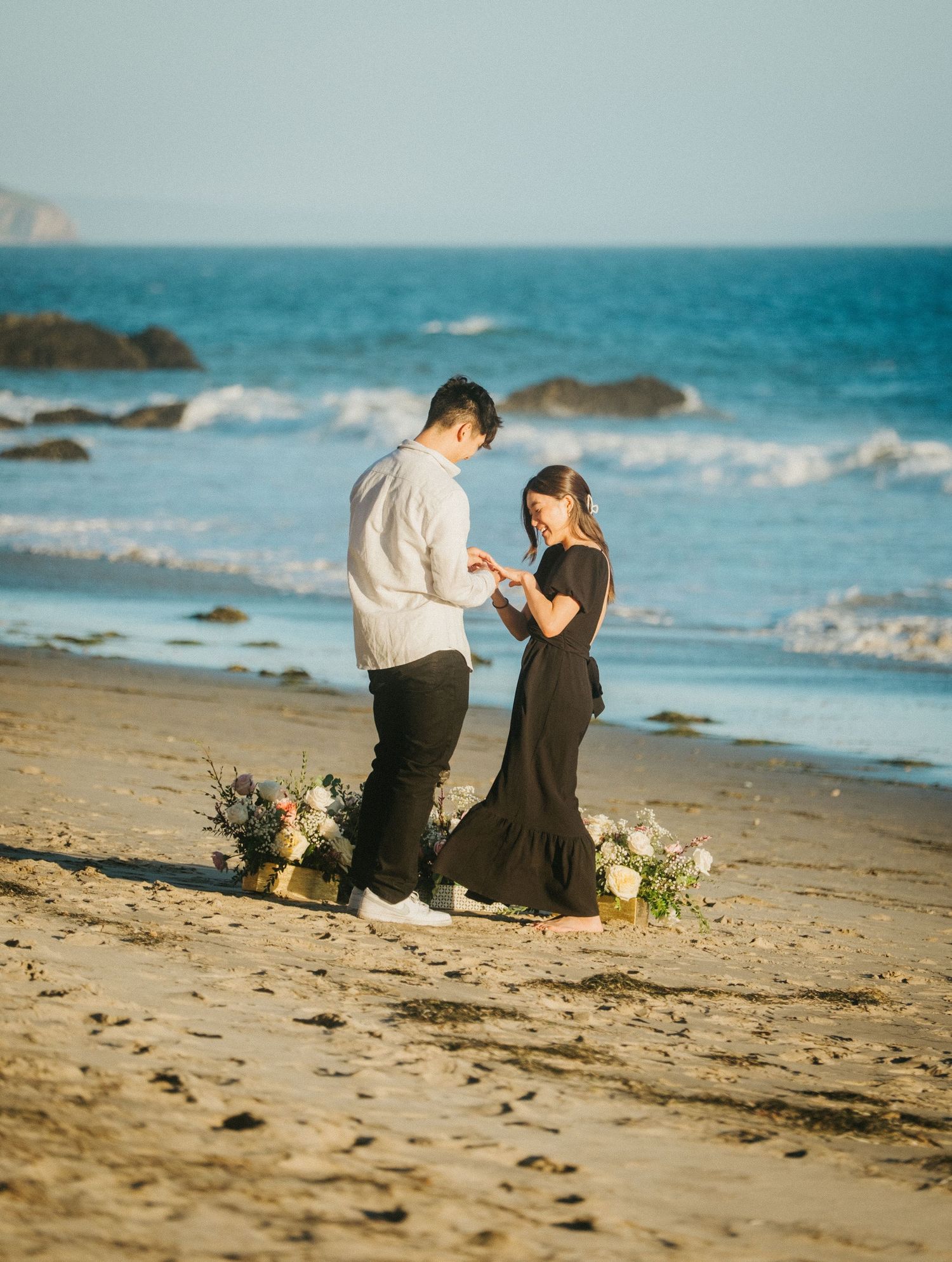 Couple exchanging rings during beach proposal with floral arrangements nearby.