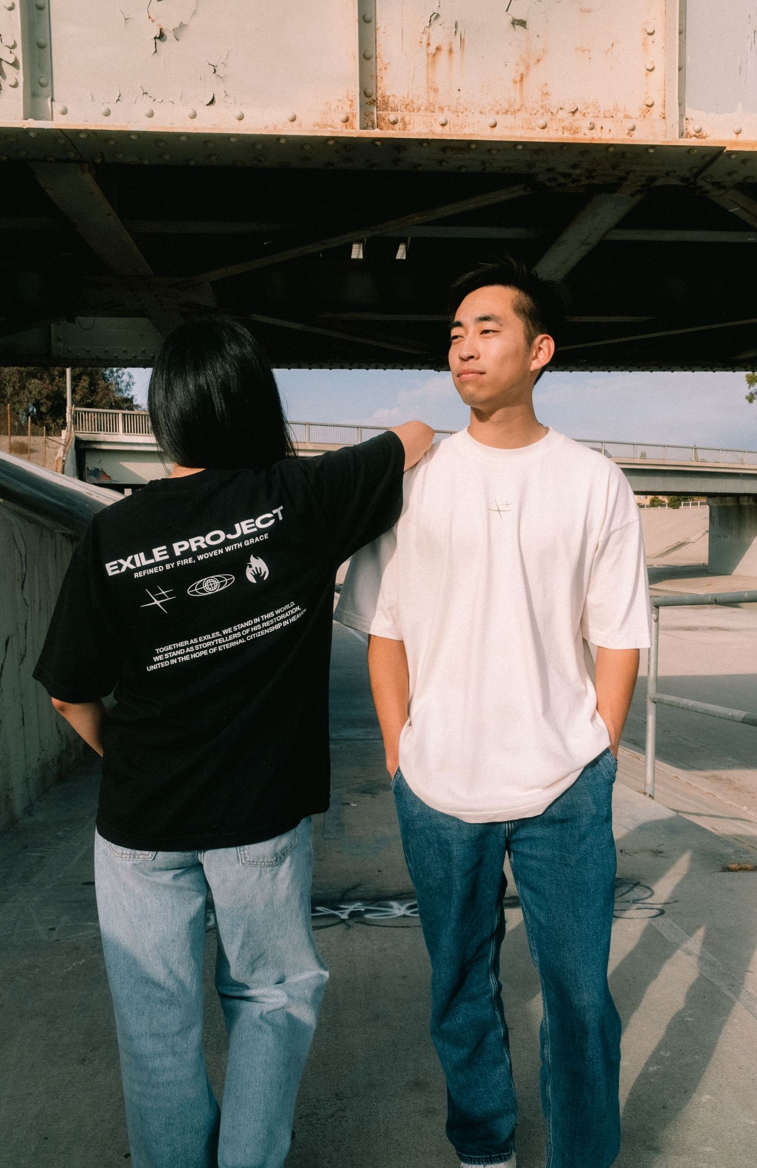 Two people wearing Exile Project t-shirts in black and white under an urban overpass.
