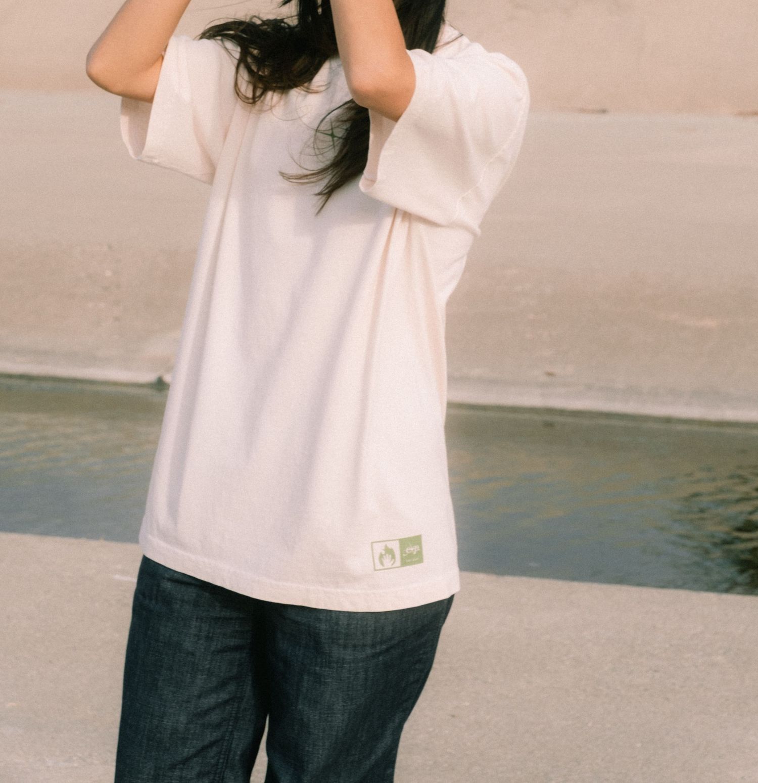 Woman wearing a white oversized t-shirt with green logo patch and dark jeans, standing outdoors.