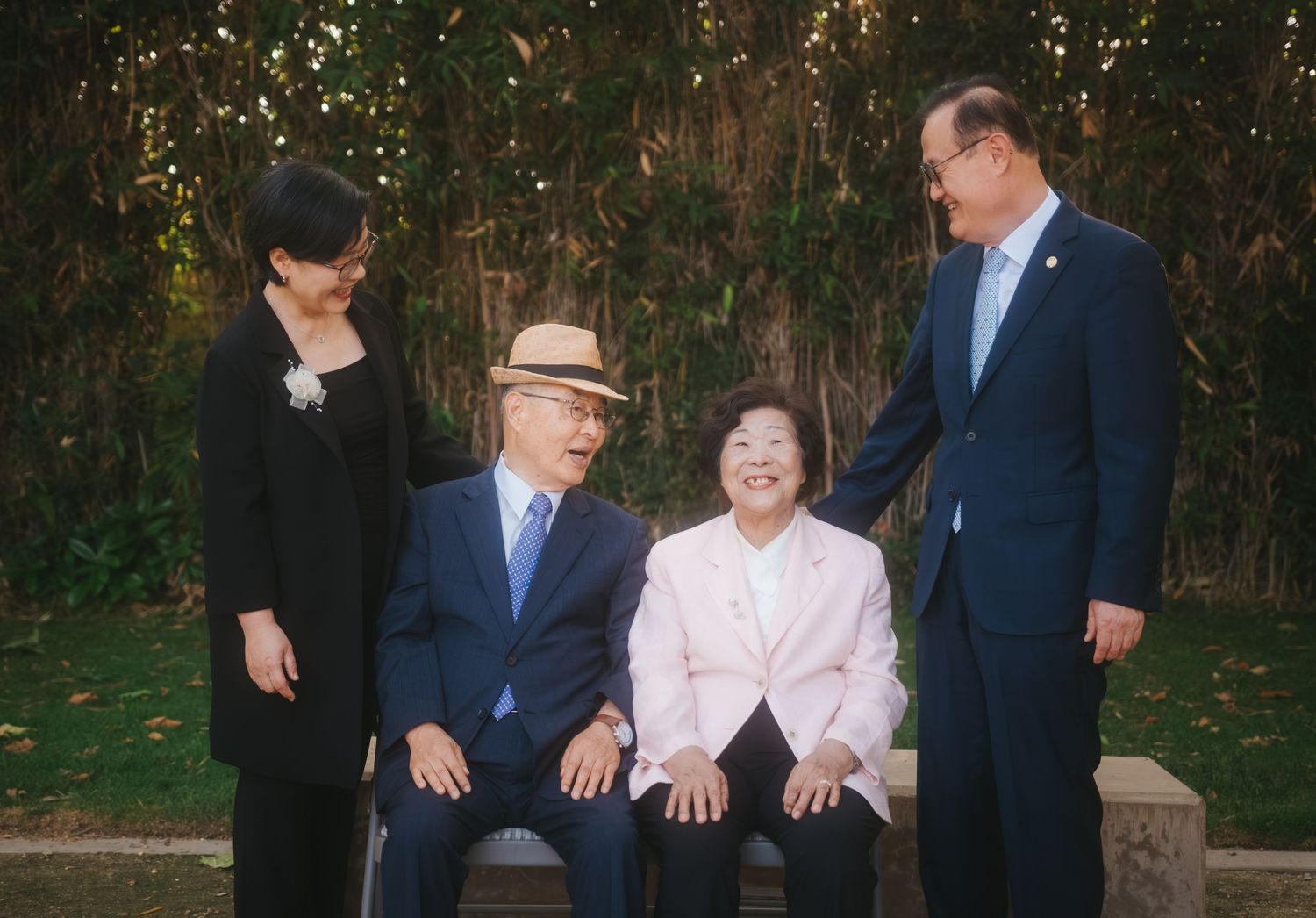 Two elderly people sit on a bench while two adults stand beside them, posing together in a lush garden setting.