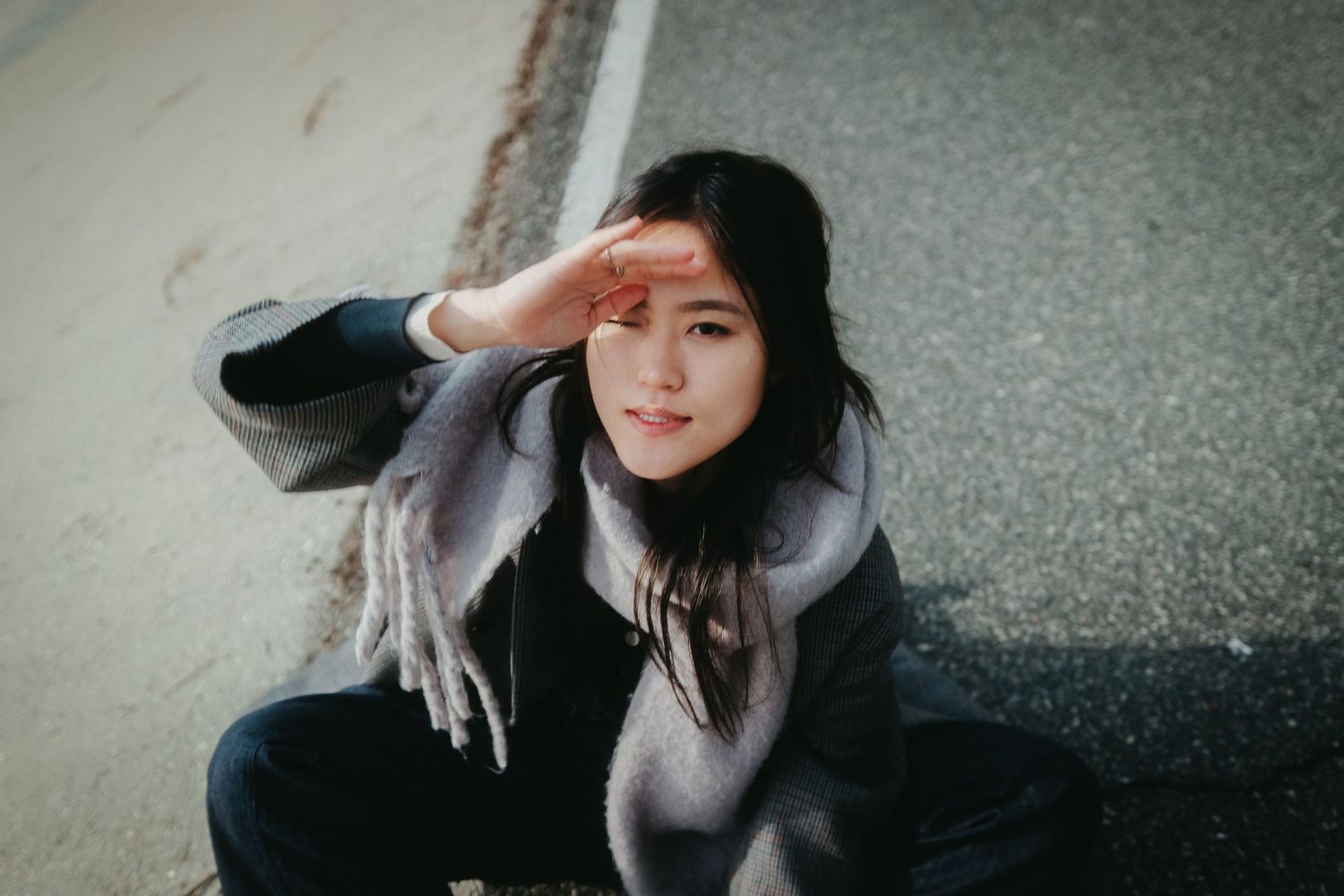 Young Asian woman sitting on road, shielding eyes from sun with hand, wearing gray scarf and dark jacket.