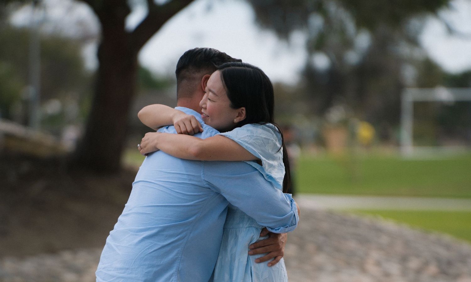 Two people sharing an emotional hug outdoors in a park, wearing matching light blue shirts on a sunny day.