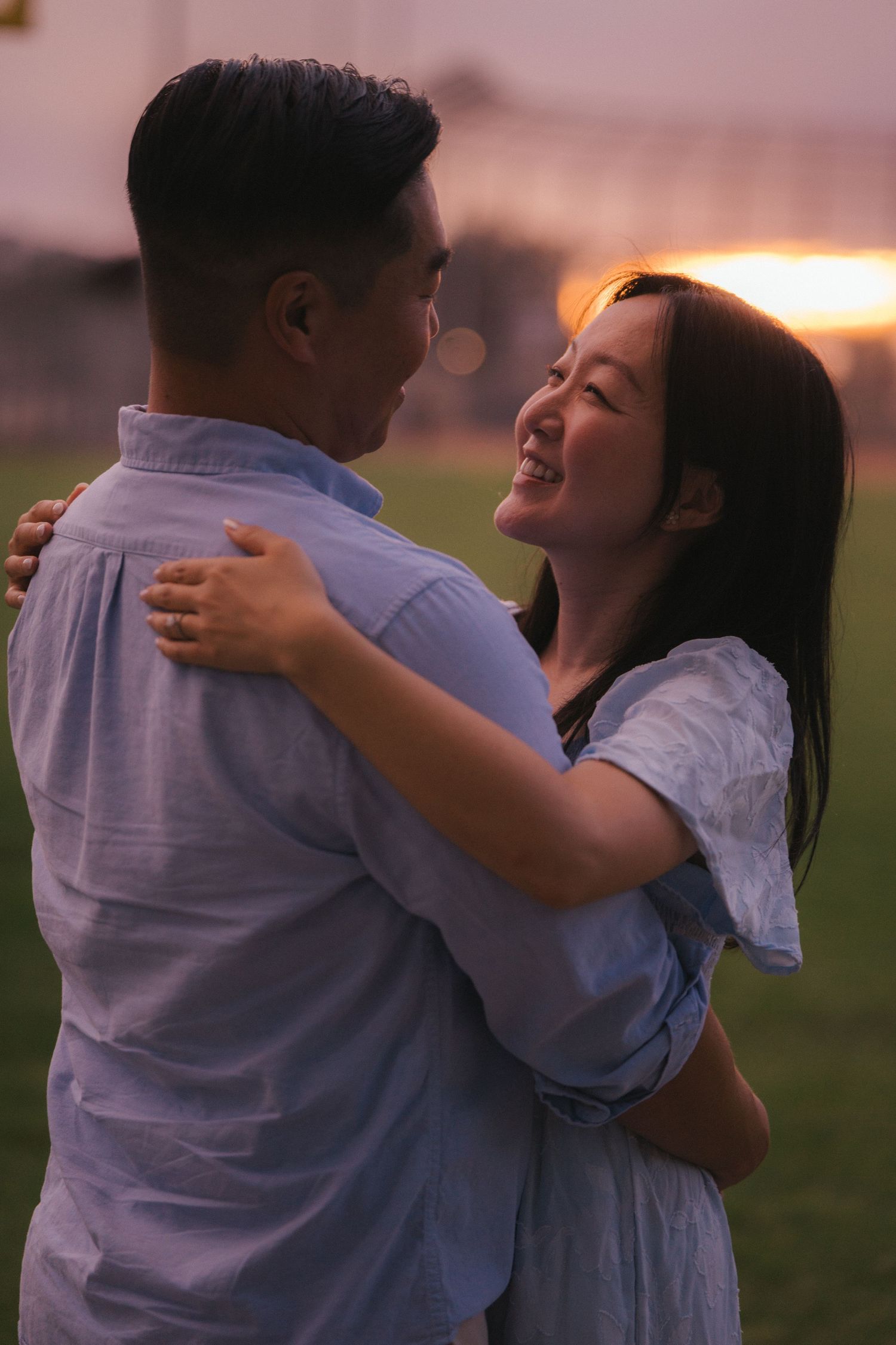 Romantic Asian couple holding each other close in park field during golden sunset, woman smiling.