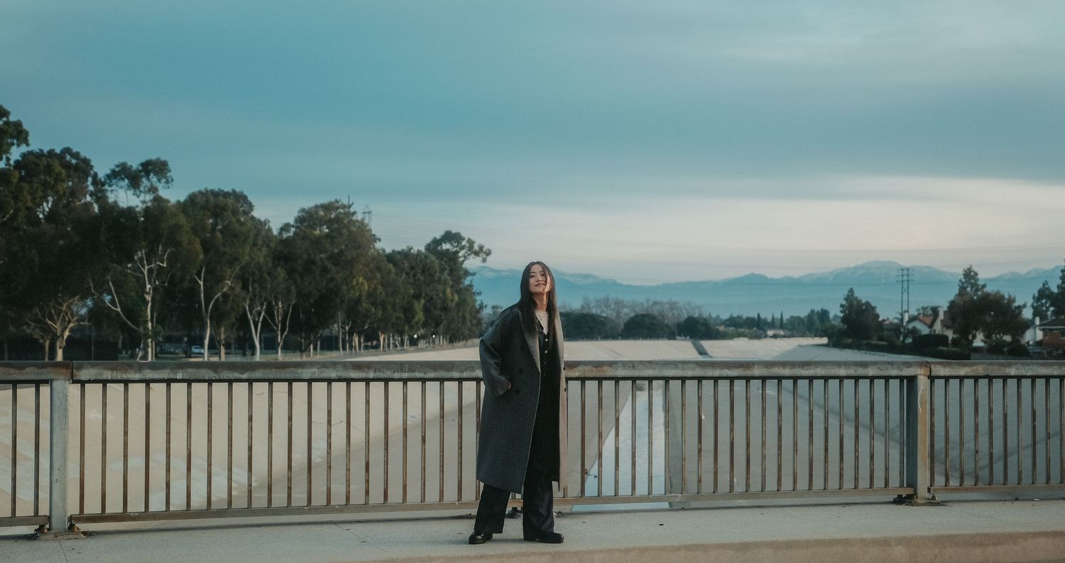 Woman in black coat standing on bridge overlooking serene lake with mountain backdrop and lush trees.