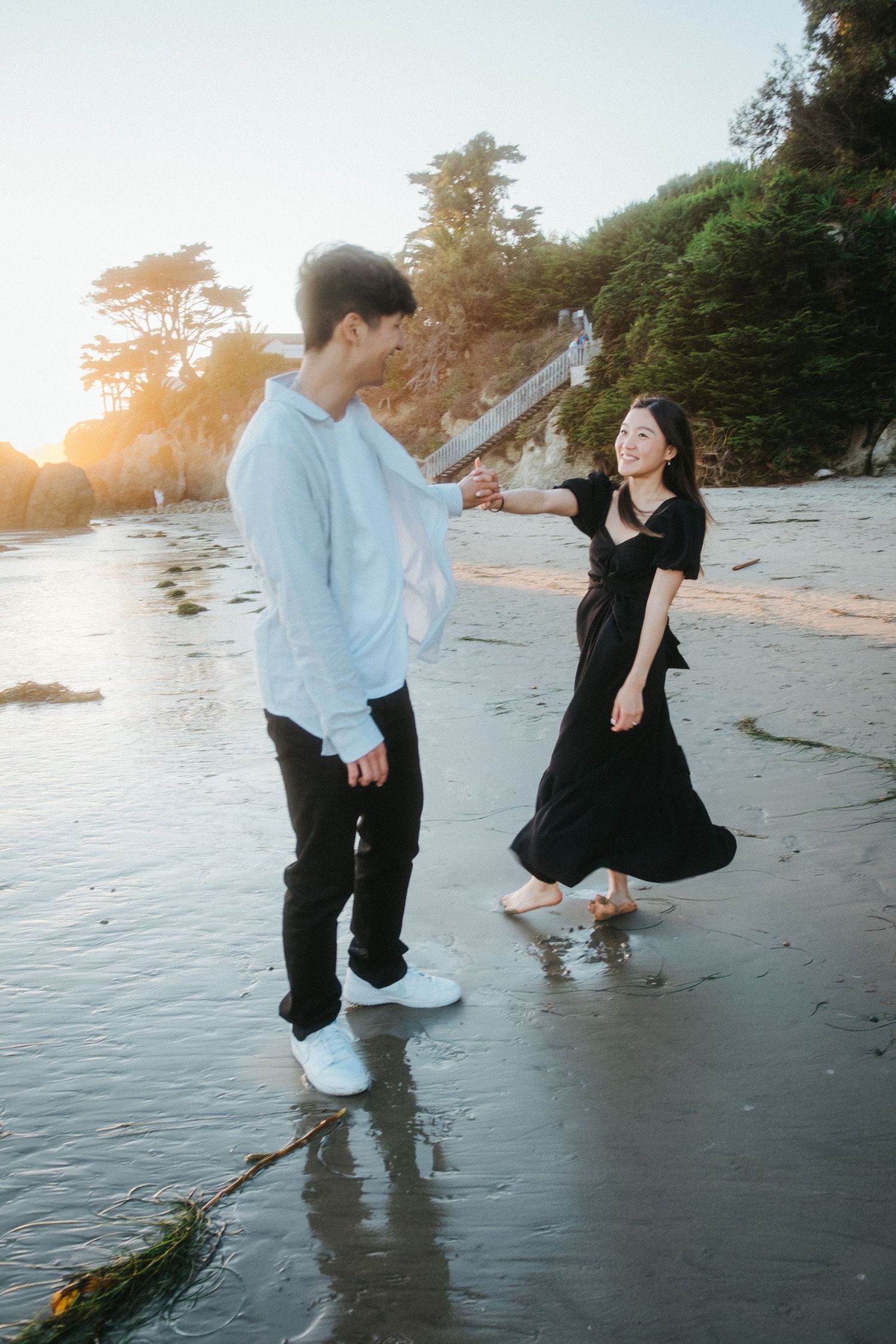 Young couple playfully dancing on a wet beach at golden hour, woman in black dress laughing and spinning.