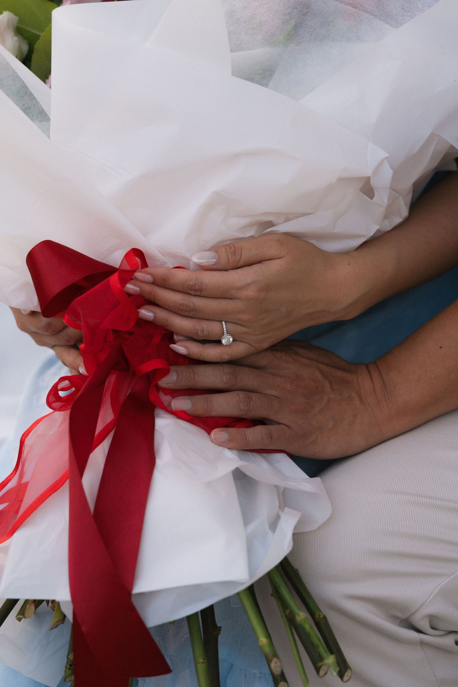 Close-up of engaged couple's hands holding white bouquet with red ribbon, diamond engagement ring visible.