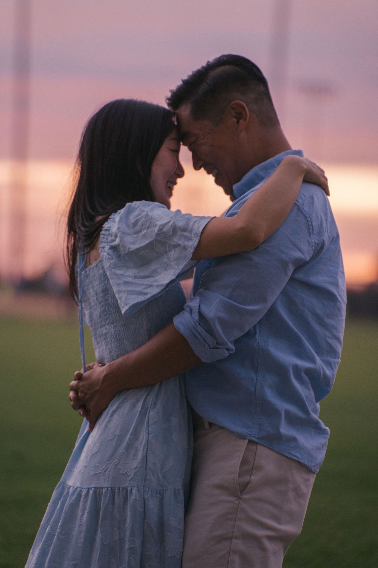 Tender couple touching foreheads in intimate embrace on grass field during purple sunset twilight.