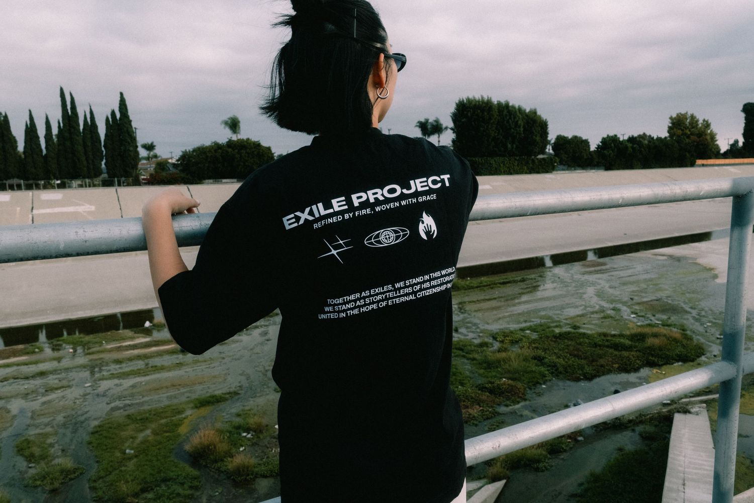 Person wearing black Exile Project graphic t-shirt standing on a bridge over water with trees in background.