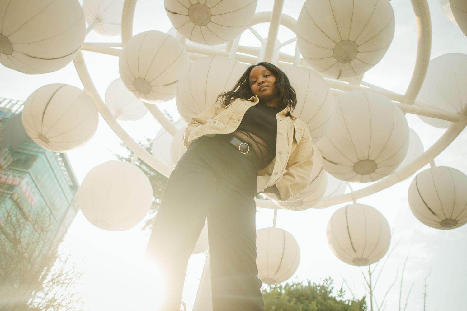 Woman in beige jacket posing beneath white paper lanterns in a bright outdoor art installation.