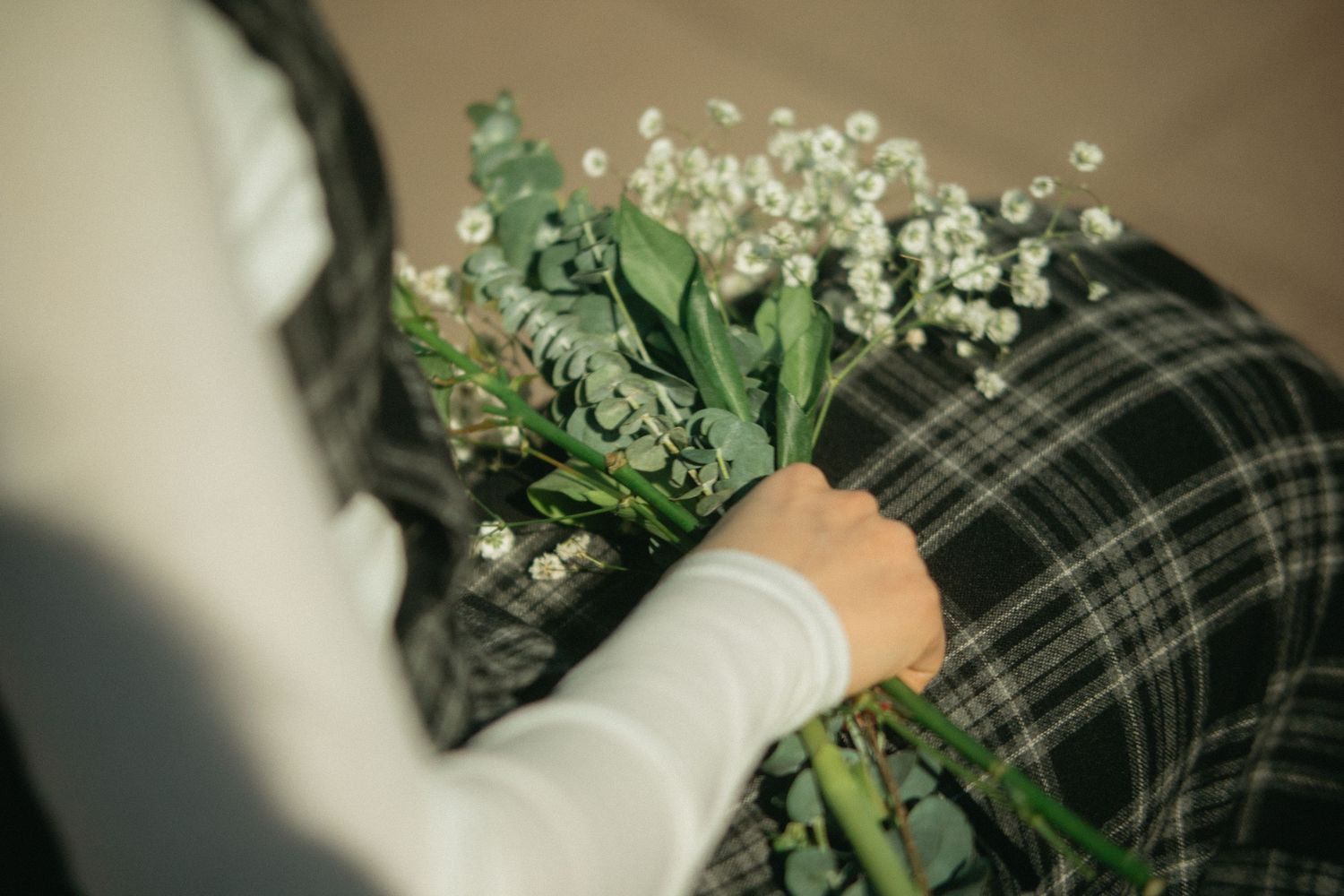 Close-up of hands holding a bouquet of white flowers and eucalyptus on a plaid skirt.