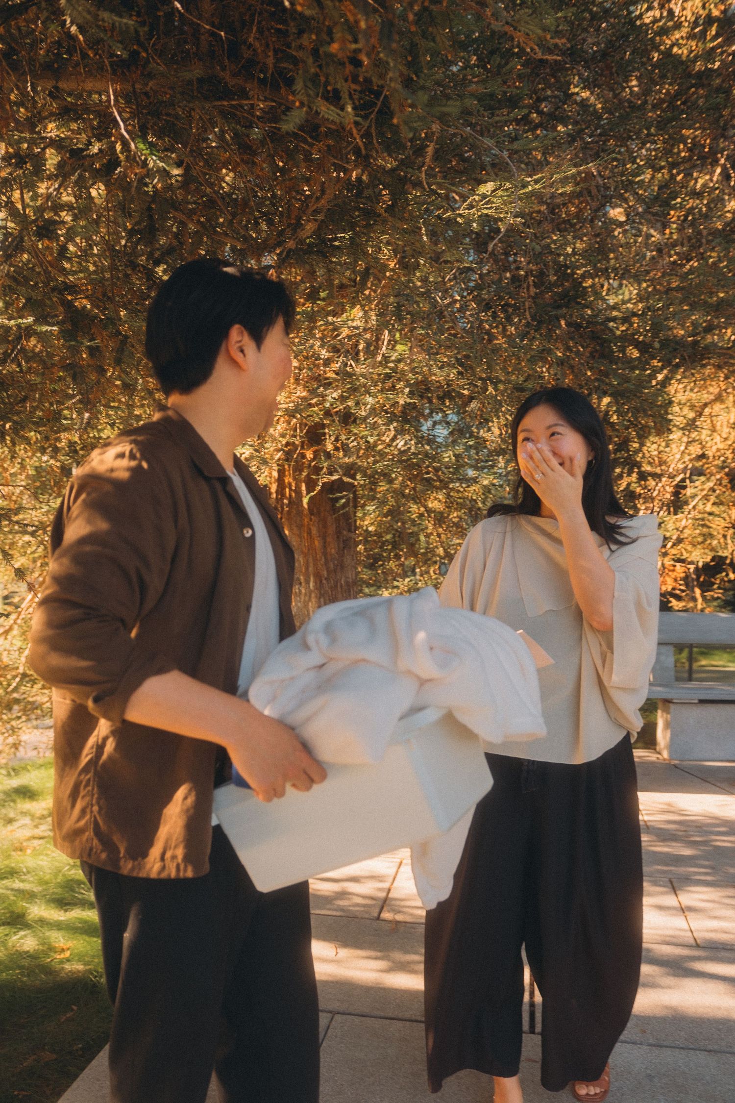 Asian man surprises woman with a wrapped gift outdoors, as she covers her mouth in delight.