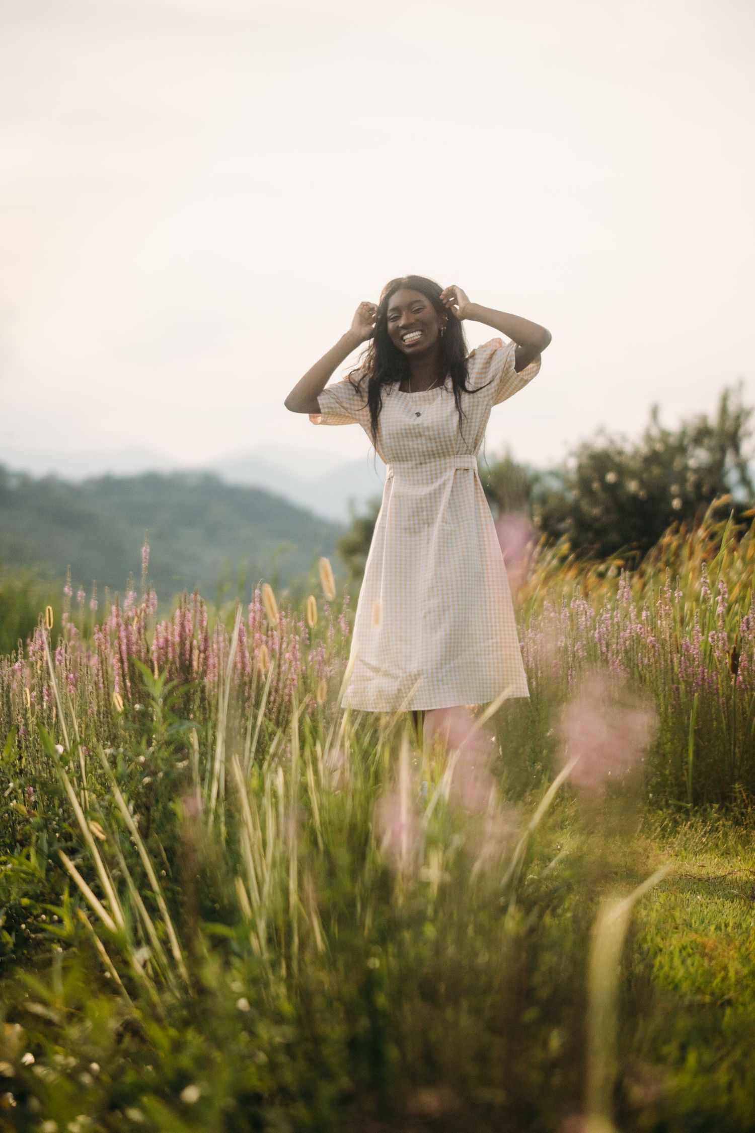 A joyful Black woman in a white dress laughs while touching her hair in a field of purple wildflowers.