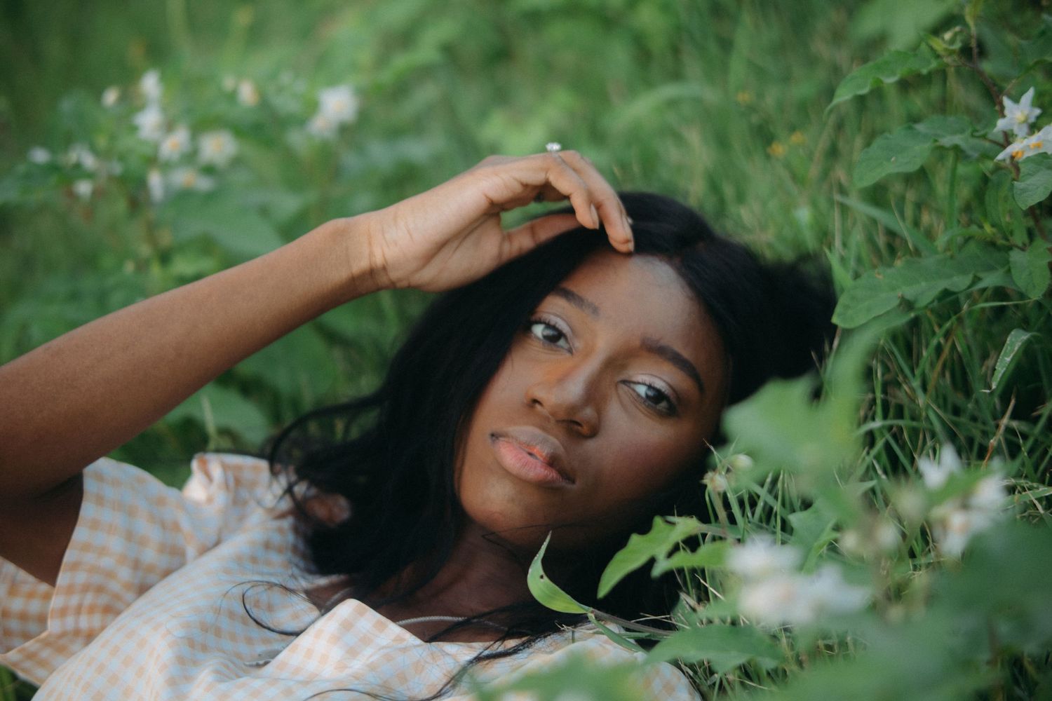 Young Black woman with long hair resting among green foliage and white wildflowers in a serene outdoor setting.