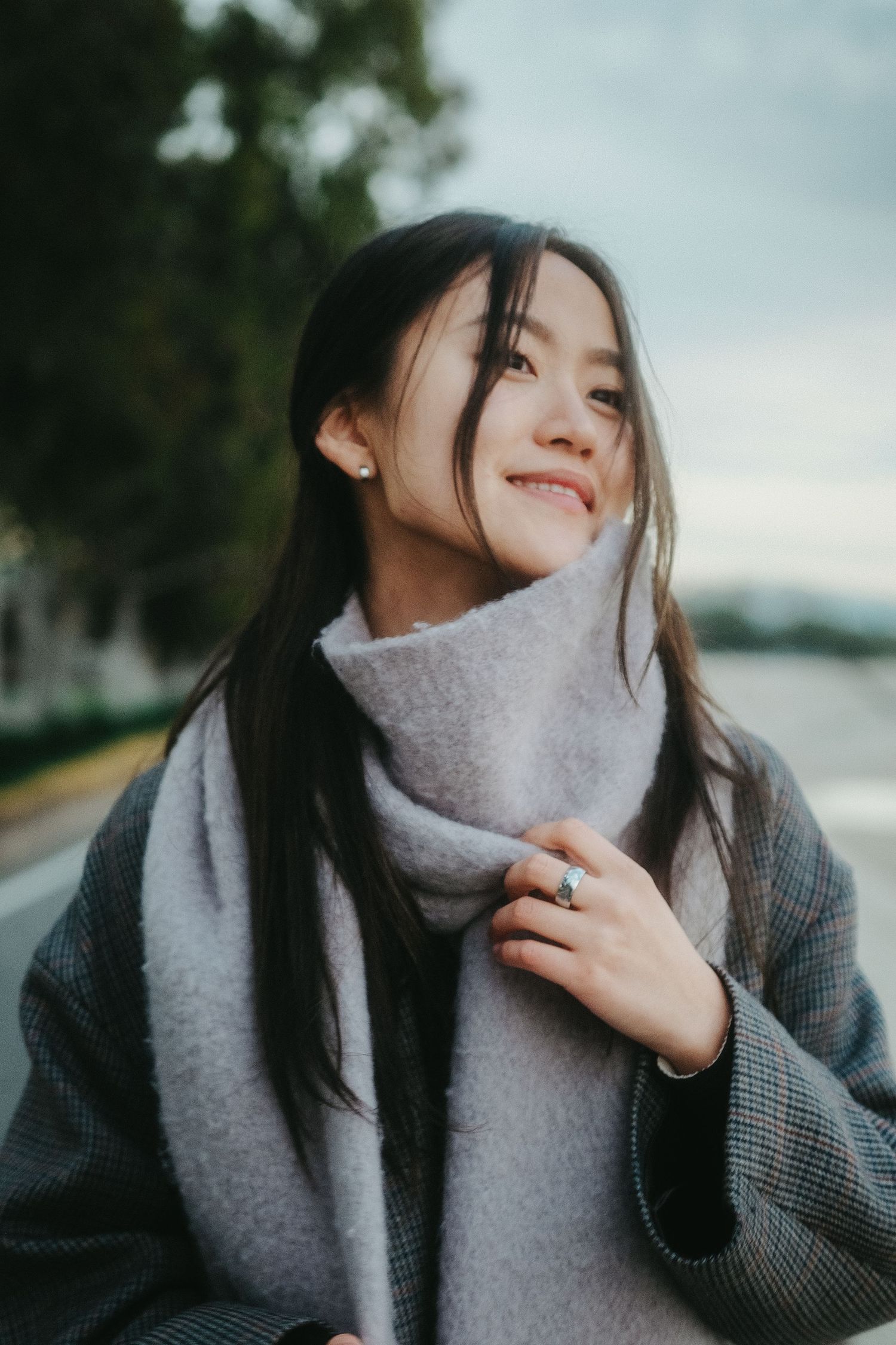Smiling young Asian woman with long dark hair wearing a gray coat and scarf, holding it close outdoors.