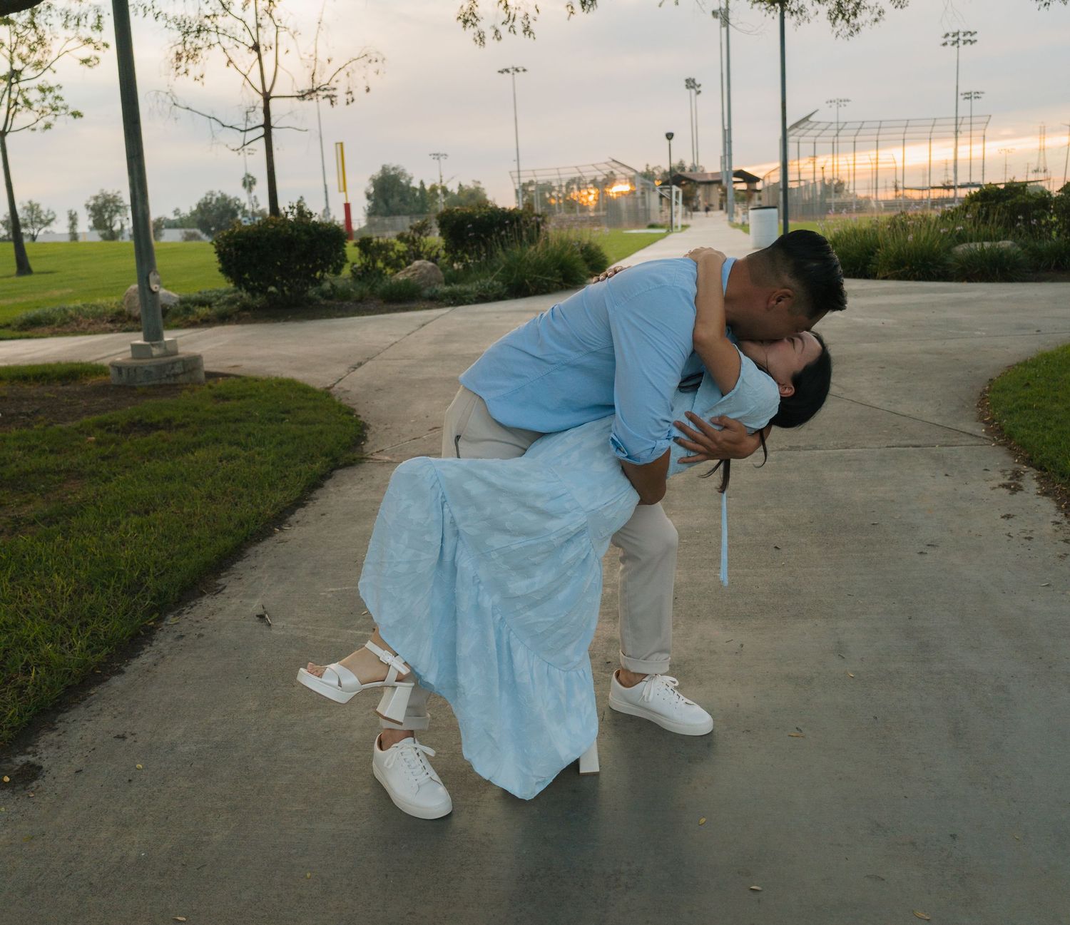 Couple sharing a romantic dip kiss on a park pathway at dusk, wearing light blue outfits and white sneakers.