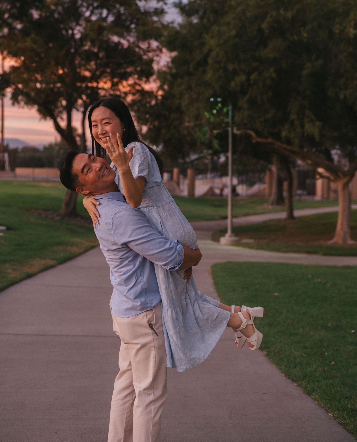 Joyful couple celebrating engagement at park during sunset, man lifting woman as she shows ring.