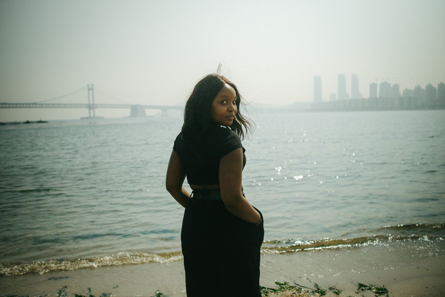 Woman in black dress standing on a beach looking back at camera with city skyline behind her.