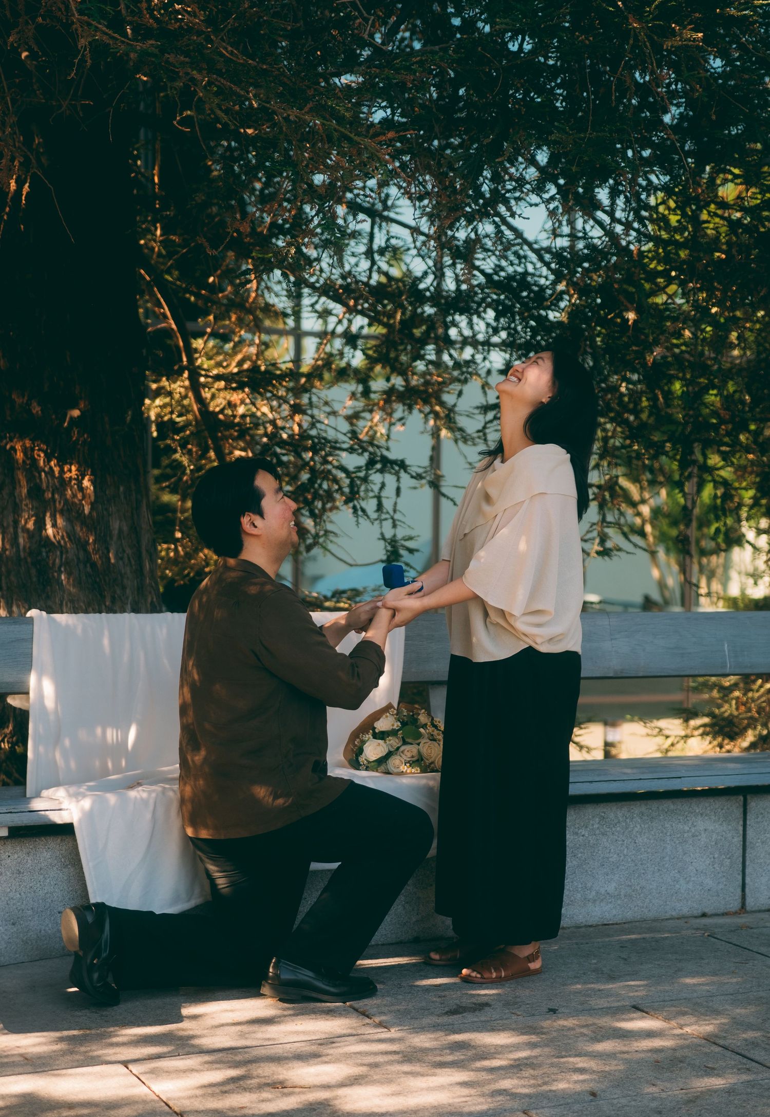 Man kneeling on one knee proposing to a woman outdoors under sunlit autumn trees.